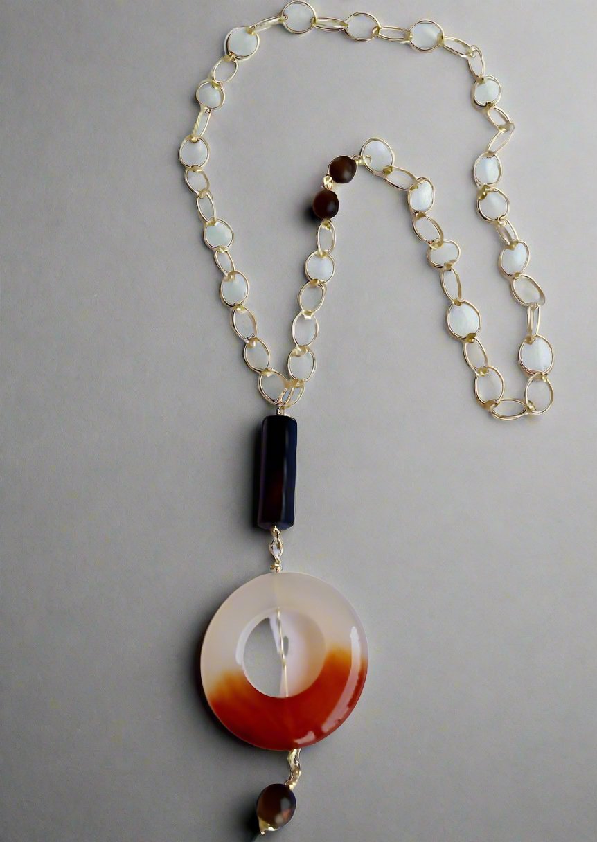 The Ariaba Onyx, Moonstone and Agate Drop Necklace by bibianna.co.uk features a gold chain with white round beads, a striking brown and white gradient pendant, a dark rectangular bead, and small brown beads, showcased on a gray background.