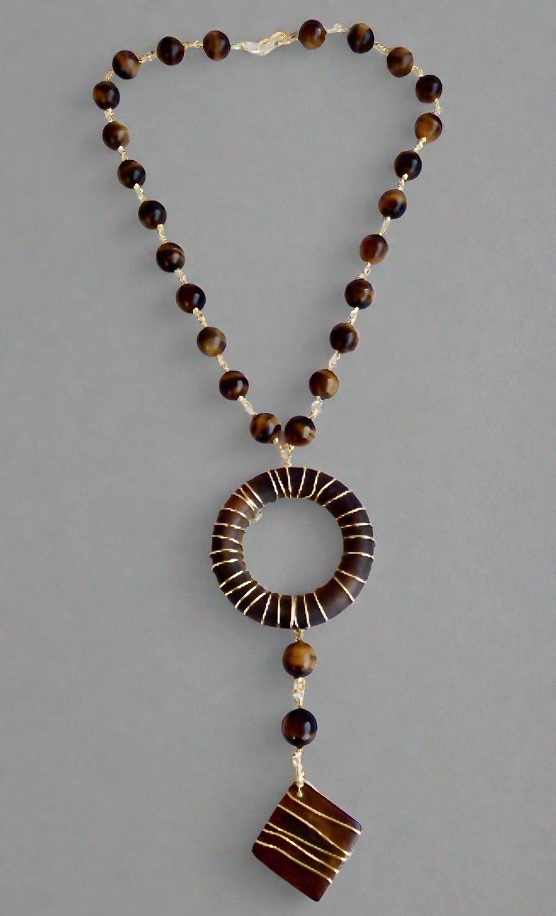 The Ariaba Wood and Tigereye Drop Necklace by bibianna.co.uk features brown and gold beads, a large circular pendant wrapped with 14k gold-plated wire, and a dangling square charm, set against a plain gray background.