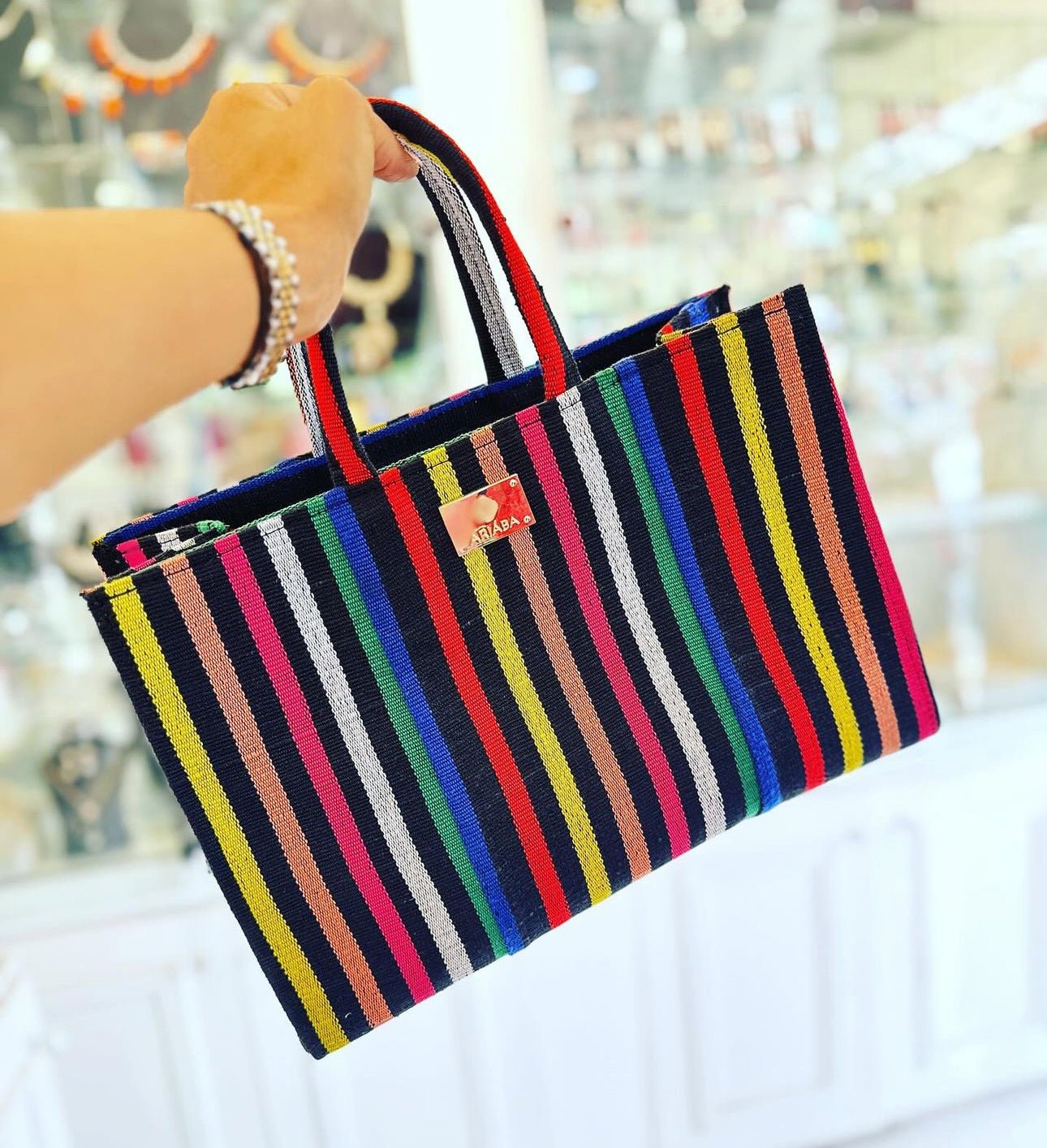 A hand holds the Ashoké Book Tote Bag - Multi Black by bibianna.co.uk, featuring colorful vertical stripes and Ashoke fabric, displayed in a well-lit shop with softly blurred jewelry and accessories in the background.