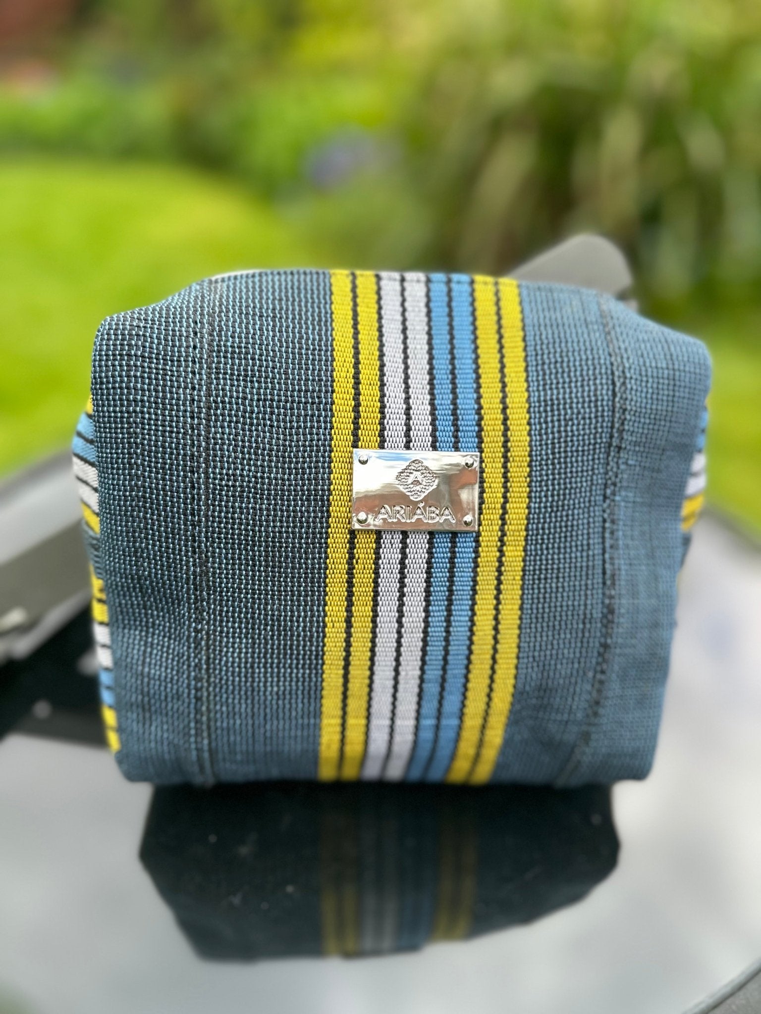 The Ashoké Book Tote Bag - Multi Blue by bibianna.co.uk, featuring blue, yellow, white, and black stripes with a metal CARLARA logo plate, is displayed on a reflective surface against a blurred green garden backdrop.