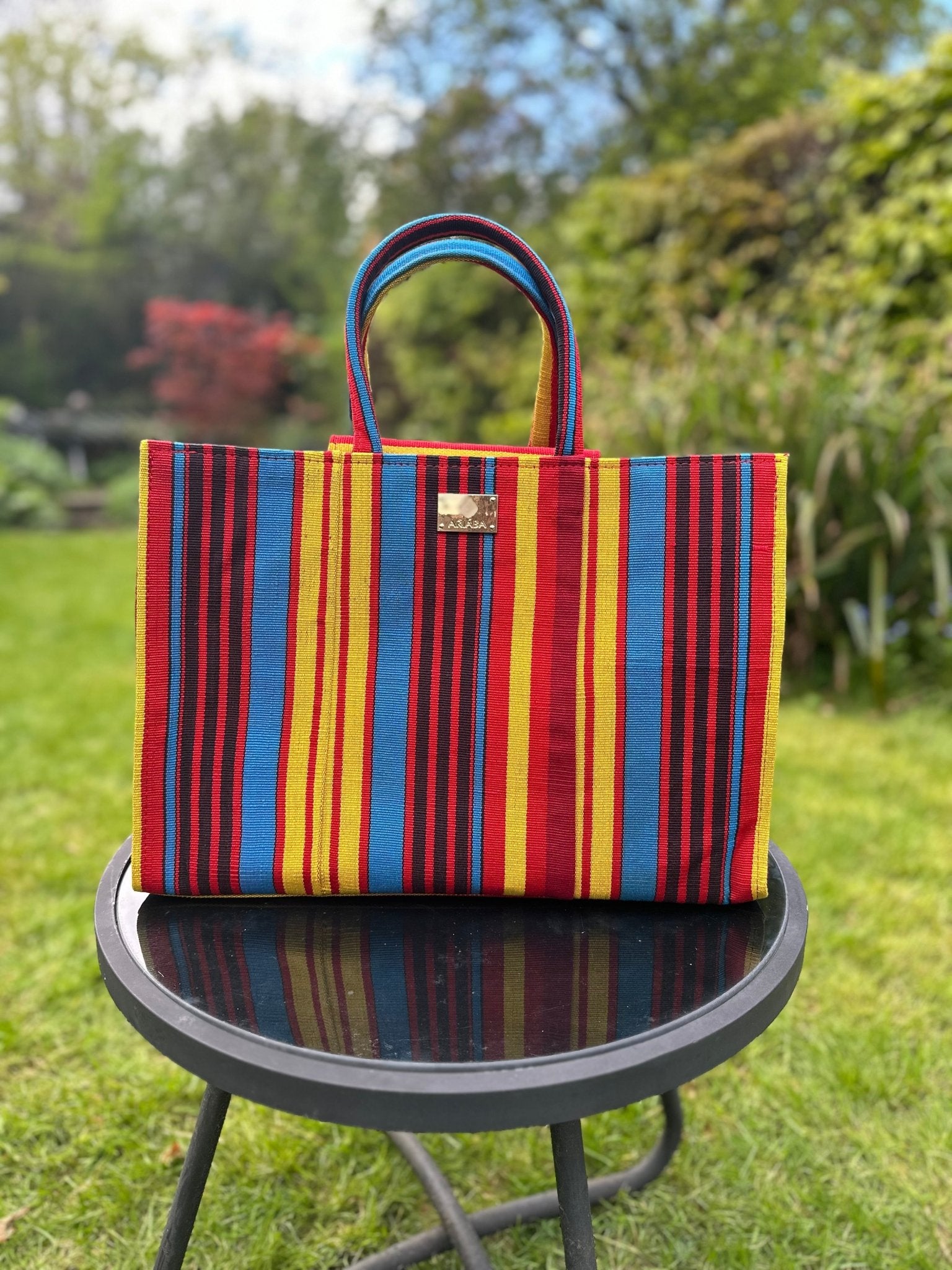 The Ashoké Book Tote Bag - Multi Red by bibianna.co.uk, handcrafted from Ashoké with bold red, yellow, blue, and black stripes, is displayed outdoors on a round glass table against a lush garden backdrop.