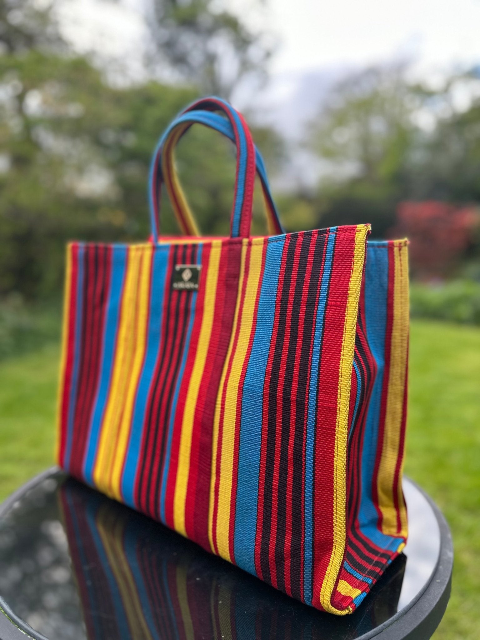 The Ashoké Book Tote Bag - Multi Red by bibianna.co.uk, featuring colorful vertical stripes, sits on a glass table outdoors with a blurred garden backdrop.