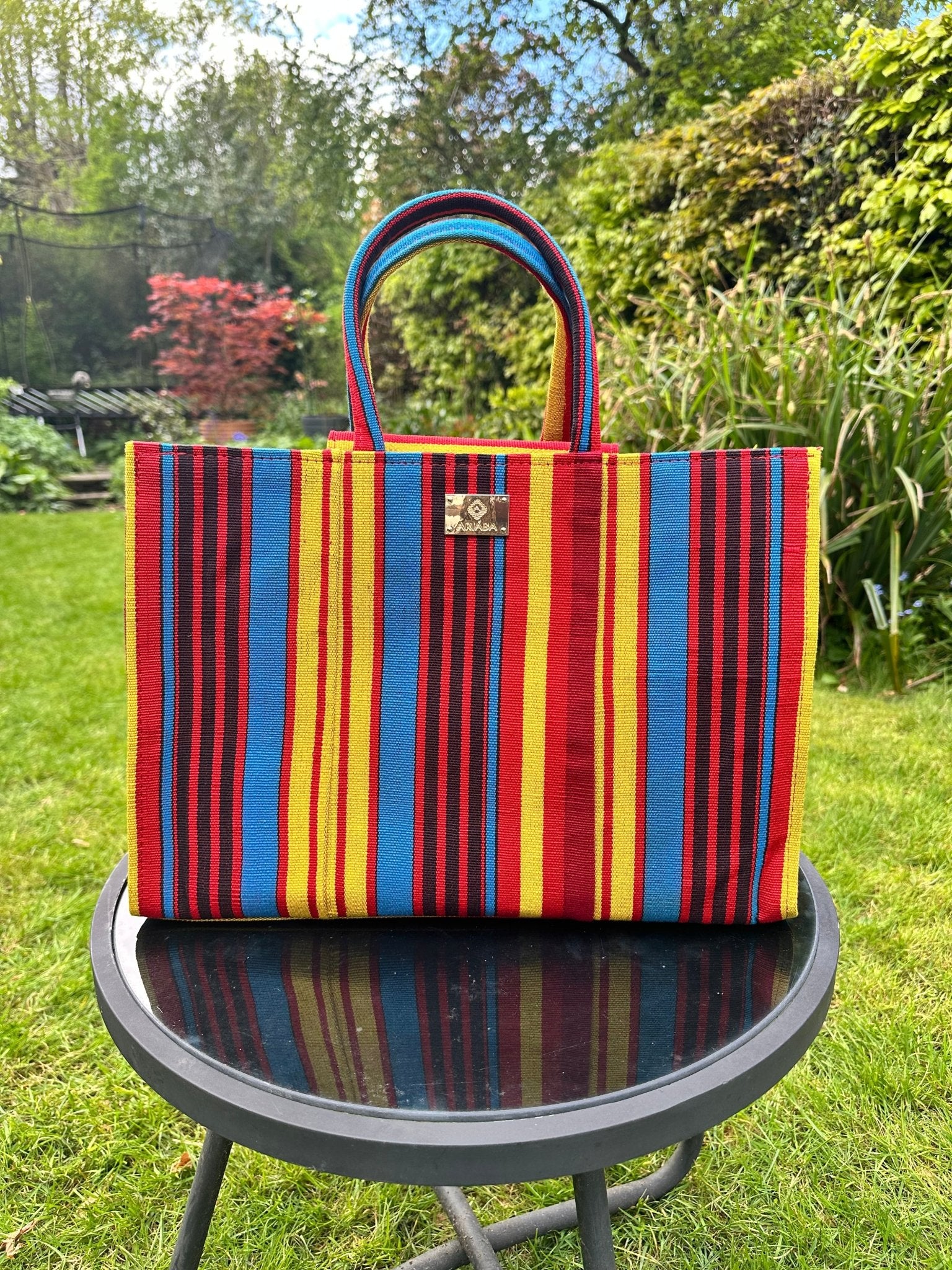 The Ashoké Book Tote Bag - Multi Red from bibianna.co.uk, handcrafted by a Nigerian designer, rests on a round glass table outdoors amid lush green grass and trees.