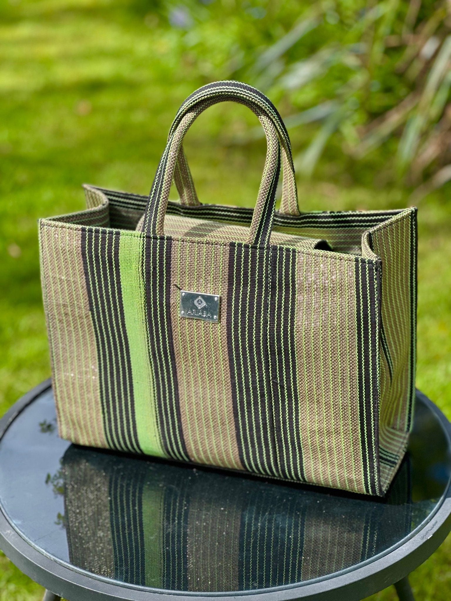 The Ashoké Book Tote Bag - Multi Sage by bibianna.co.uk, featuring green, brown, and black Ashoké stripes, sits on a round glass table outdoors amid lush grass and plants.