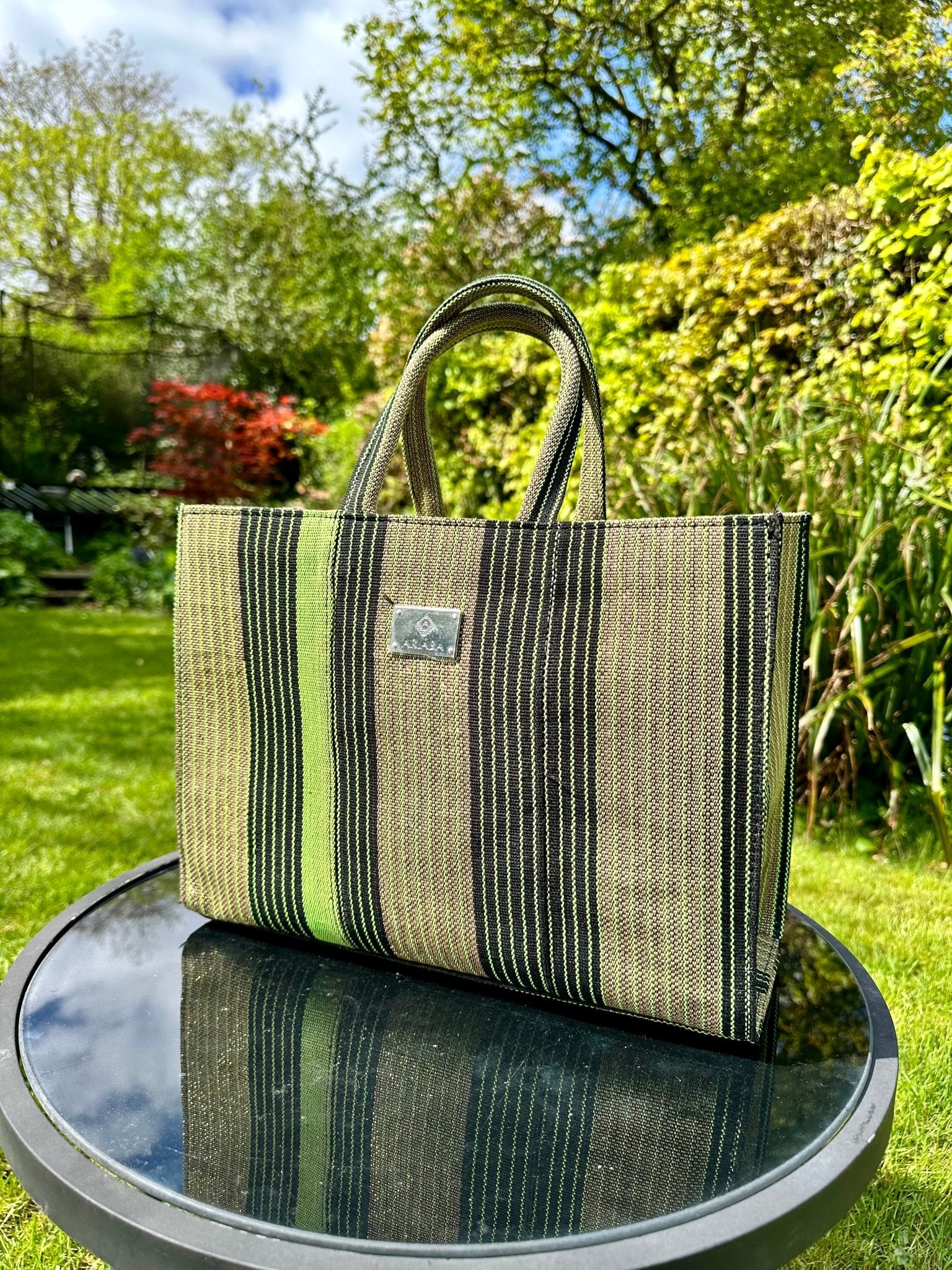The Ashoké Book Tote Bag - Multi Sage by bibianna.co.uk, a large green and black striped tote with short handles, sits on a round glass table in a sunny garden with lush greenery and colorful plants behind it.