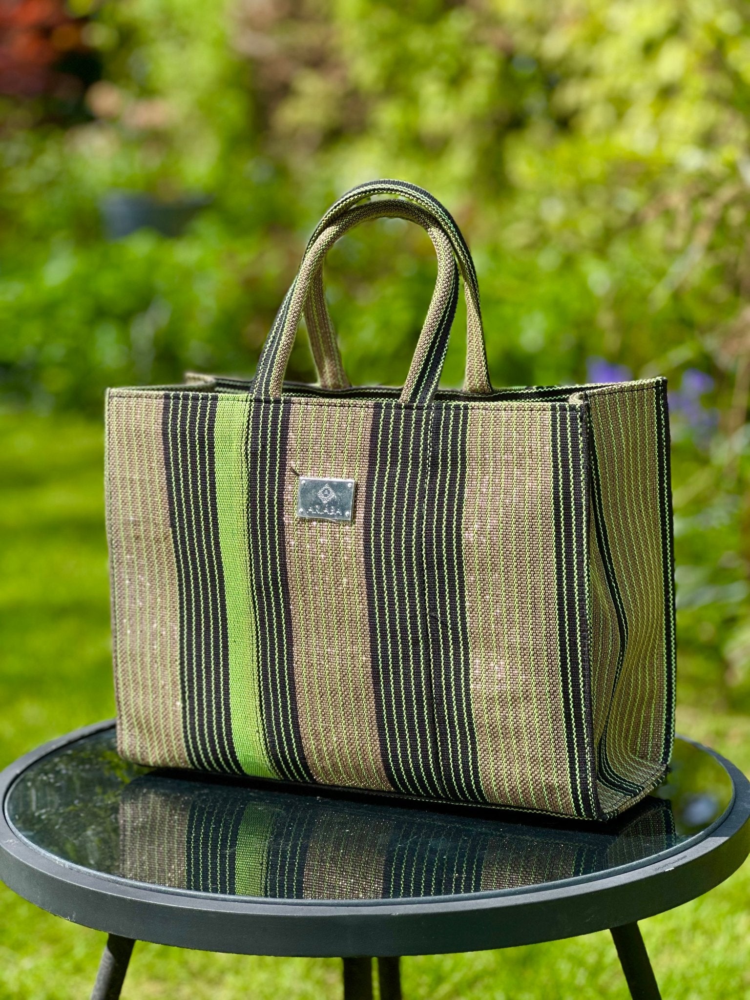 The Ashoké Book Tote Bag - Multi Sage by bibianna.co.uk, featuring green and black stripes inspired by Ashoké colours, rests on a glass-top outdoor table with a blurred garden backdrop of grass and foliage.