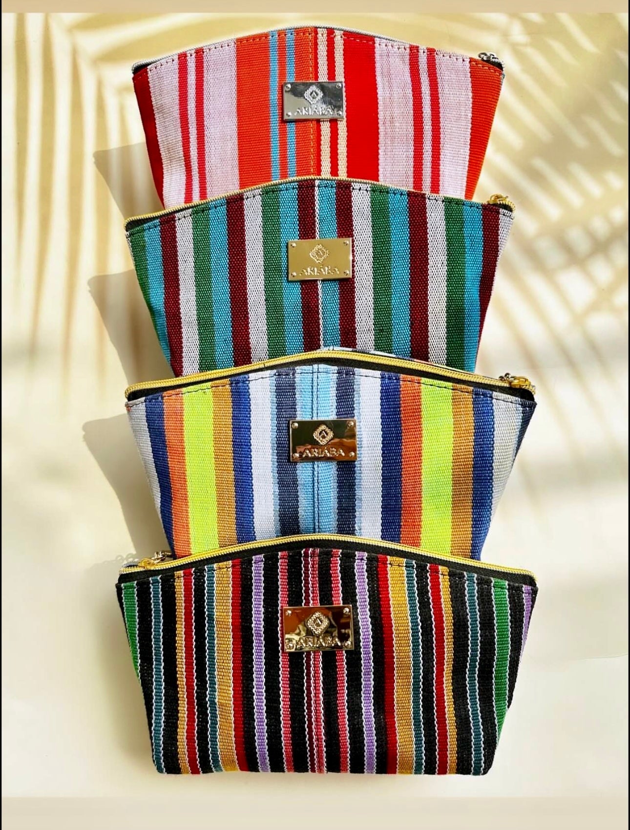 Four zippered Ashoké Purse/Makeup bags from bibianna.co.uk, each with colorful vertical stripes and gold labels, are stacked on a light background with a palm leaf shadow. Ideal as a purse or stylish makeup bag.