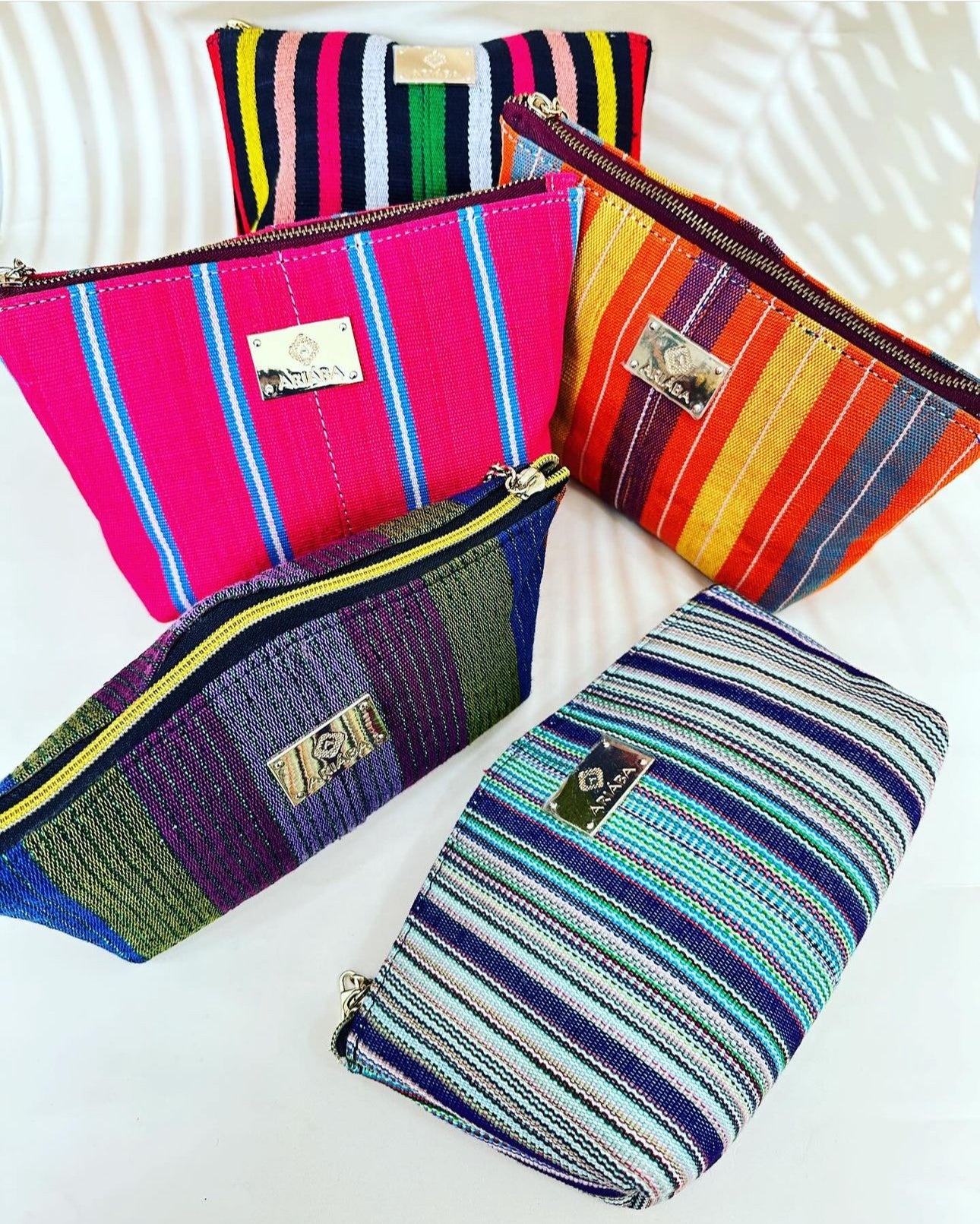 Five vibrant Ashoké Purse/Makeup bags from bibianna.co.uk, each with bold stripes and a metallic front label, are displayed on a white surface—ideal for use as stylish makeup or accessory pouches.