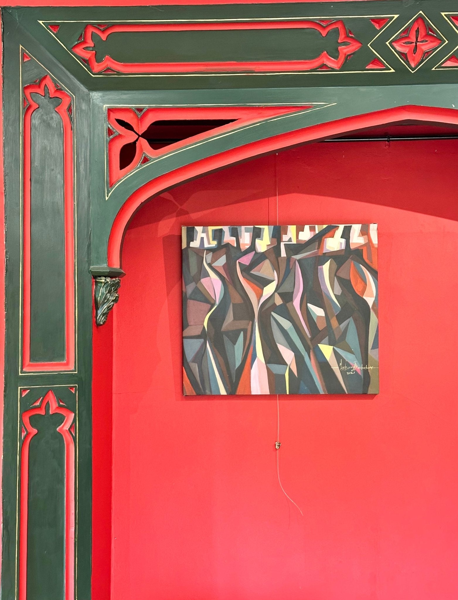 The Red Man" (78x78 cm) by Barry Ikechukwu, a vibrant mixed media acrylic piece with abstract human forms, hangs on a vivid red wall below ornate Victorian-style green and red trim with intricate cutouts. A pull string hangs beneath it.