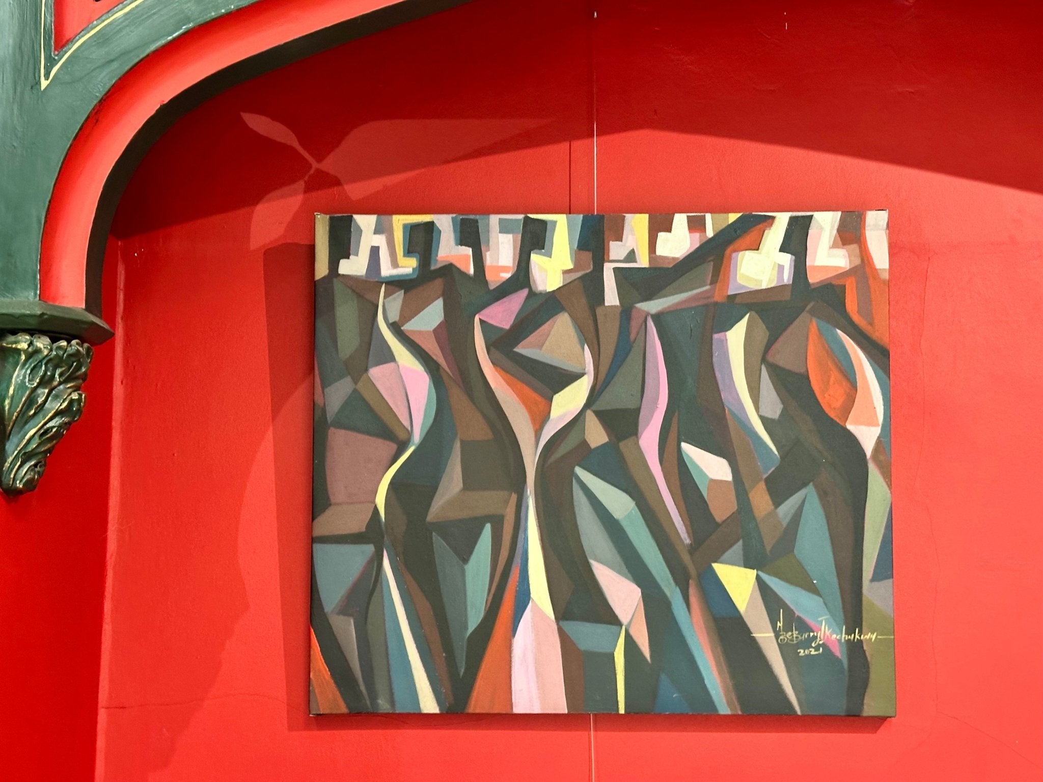 'The Red Man' by Barry Ikechukwu is a cubist abstract painting (78x78 cm) featuring geometric shapes in muted green, brown, pink, and blue tones. Shown on a red wall with signature and date visible in the bottom right corner.