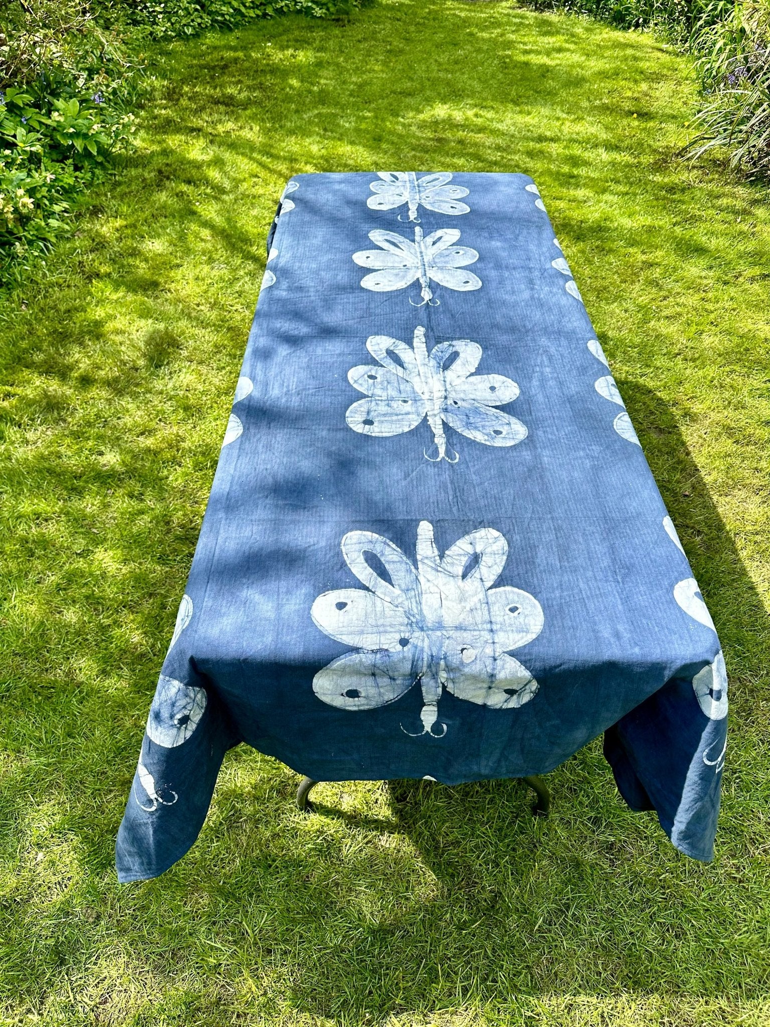 Outdoors on green grass, a long table is covered with the Batik Cotton Table Cloth - Odudu by bibianna.co.uk, featuring large white butterfly designs, surrounded by lush greenery.