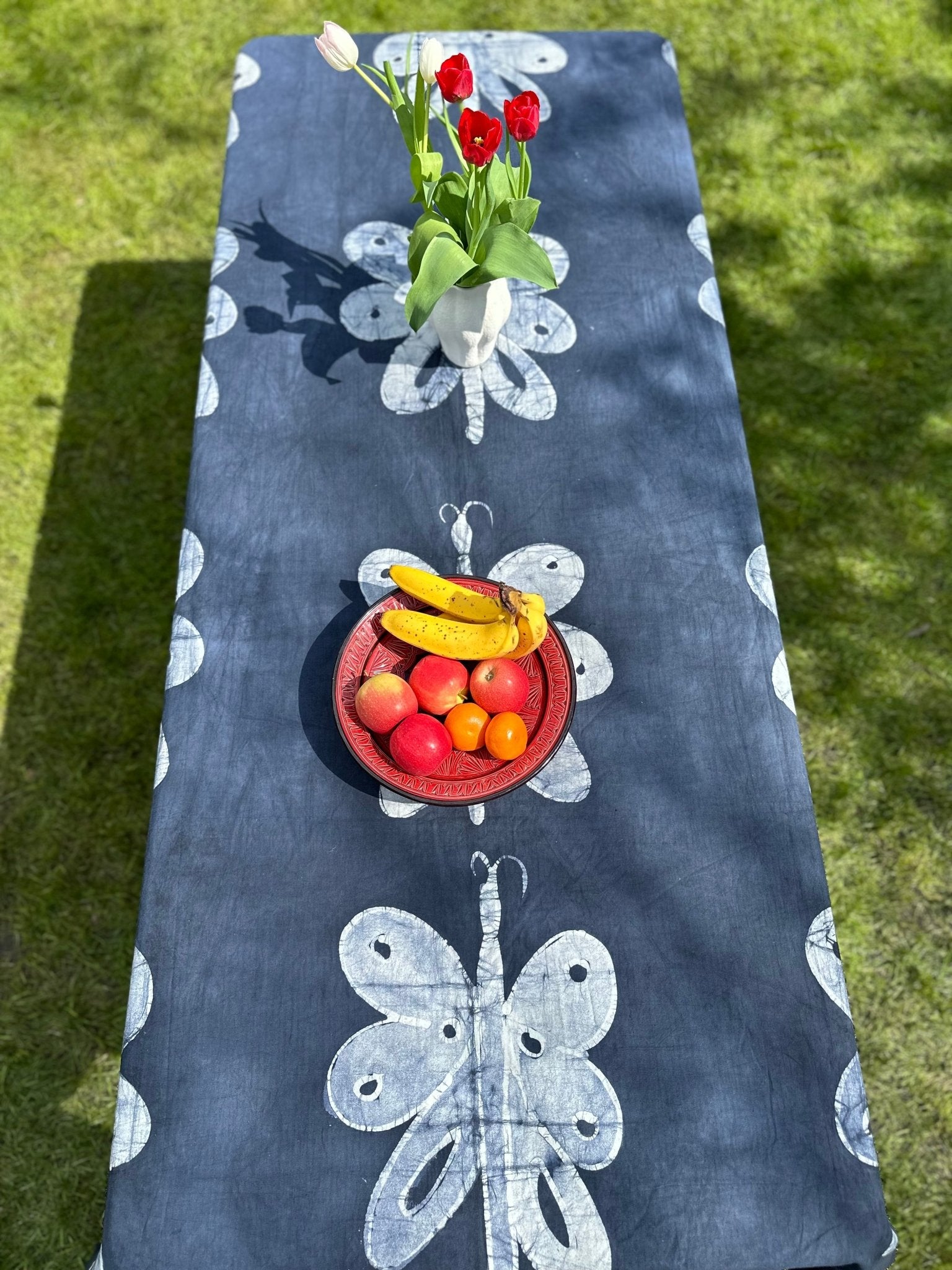 A long table draped with the bibianna.co.uk Batik Cotton Table Cloth - Odudu, handmade in Nigeria with white dragonfly patterns, stands outdoors. A vase of red tulips and a red fruit bowl decorate the tabletop.