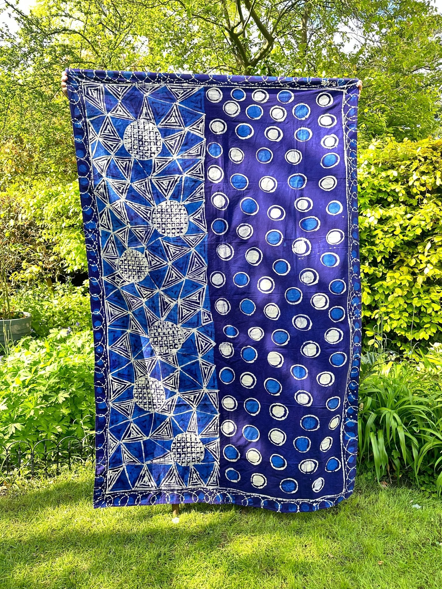 The Batik table cloth / Beach or Picnic blanket - indigo by bibianna.co.uk is a vibrant blue, handmade cotton piece from Nigeria, featuring white geometric and circular patterns with stars and grids, displayed outdoors among green foliage.