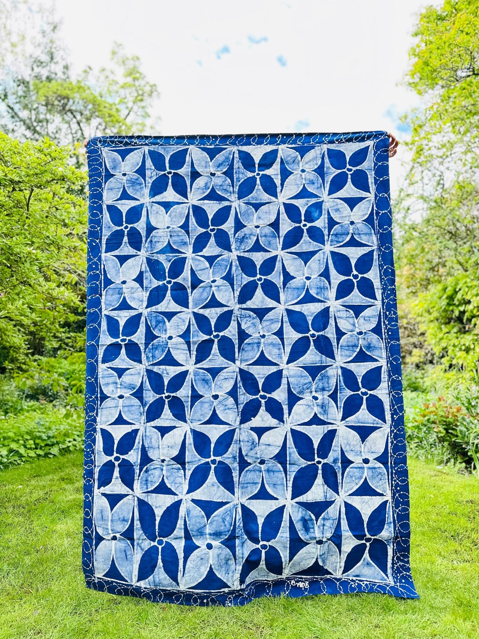 A person displays the Batik Table Cloth - Isi by bibianna.co.uk, featuring a bold blue and white circular geometric pattern, outdoors on green grass with trees in the background.