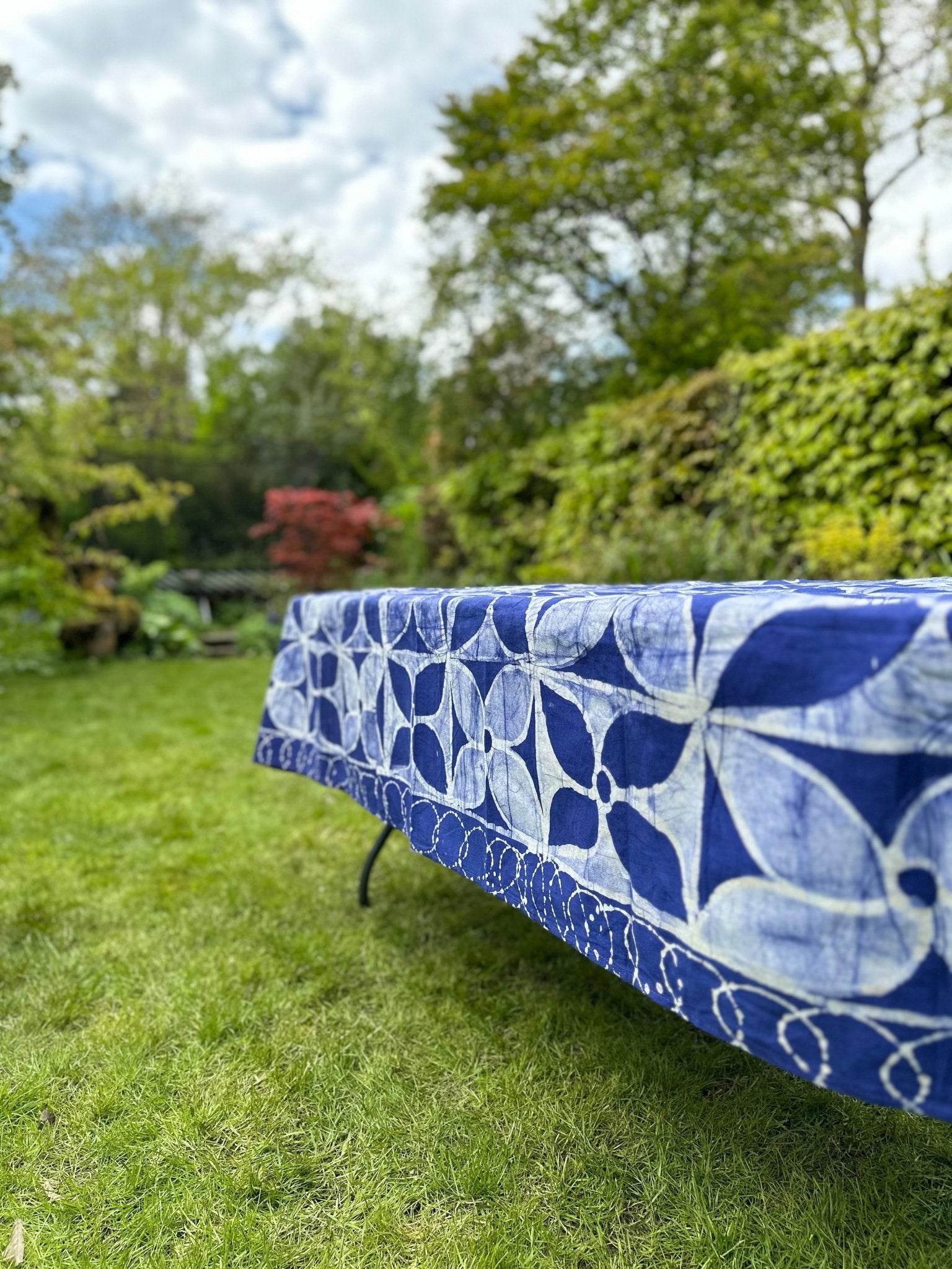 The Batik Table cloth - Isi from bibianna.co.uk, handmade by Nigerian artisans in blue and white, graces a table on green grass in a lush garden, surrounded by trees and shrubs beneath a partly cloudy sky.
