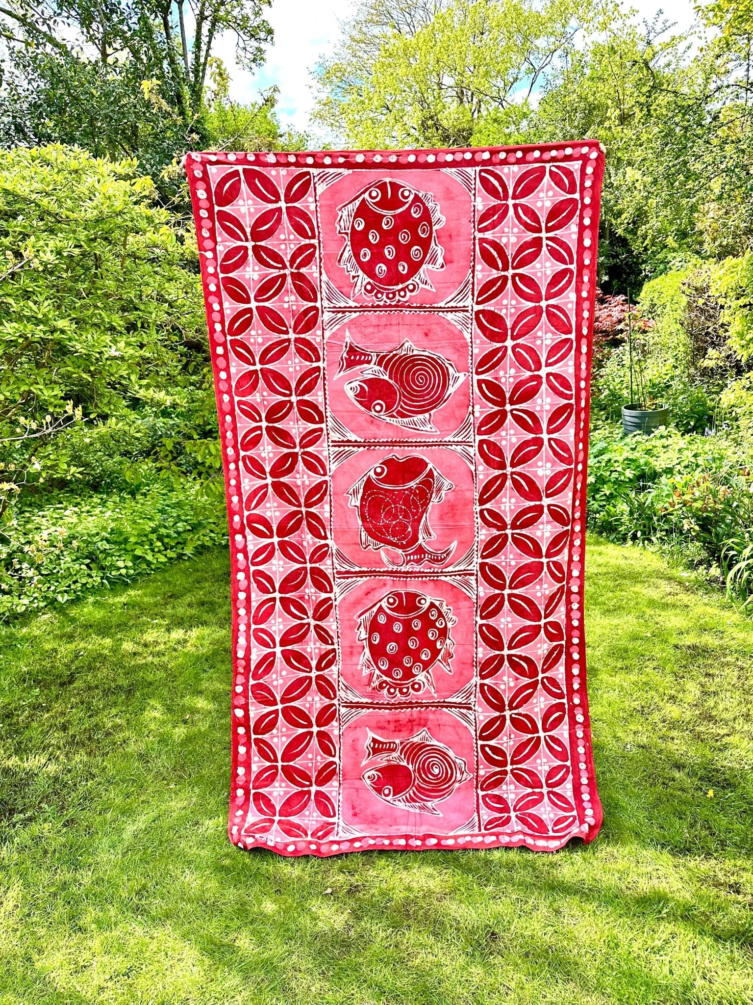 The Batik Tablecloth / Blanket - Azų Extra Large by bibianna.co.uk, handmade in Nigeria with red and white circular and spiral patterns, is displayed outdoors on a green lawn with trees and shrubs under a partly cloudy sky.