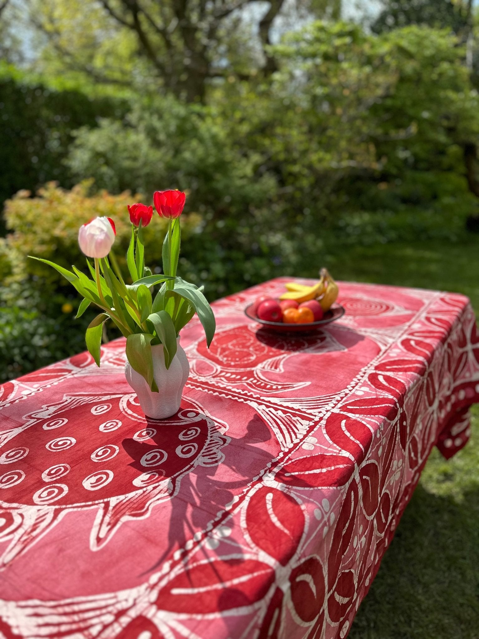 Outdoors, a Batik Tablecloth / Blanket - Azų Extra large from bibianna.co.uk covers a table topped with a white vase of red and pink tulips and a fruit bowl of bananas and apples. Lush green garden foliage fills the background.