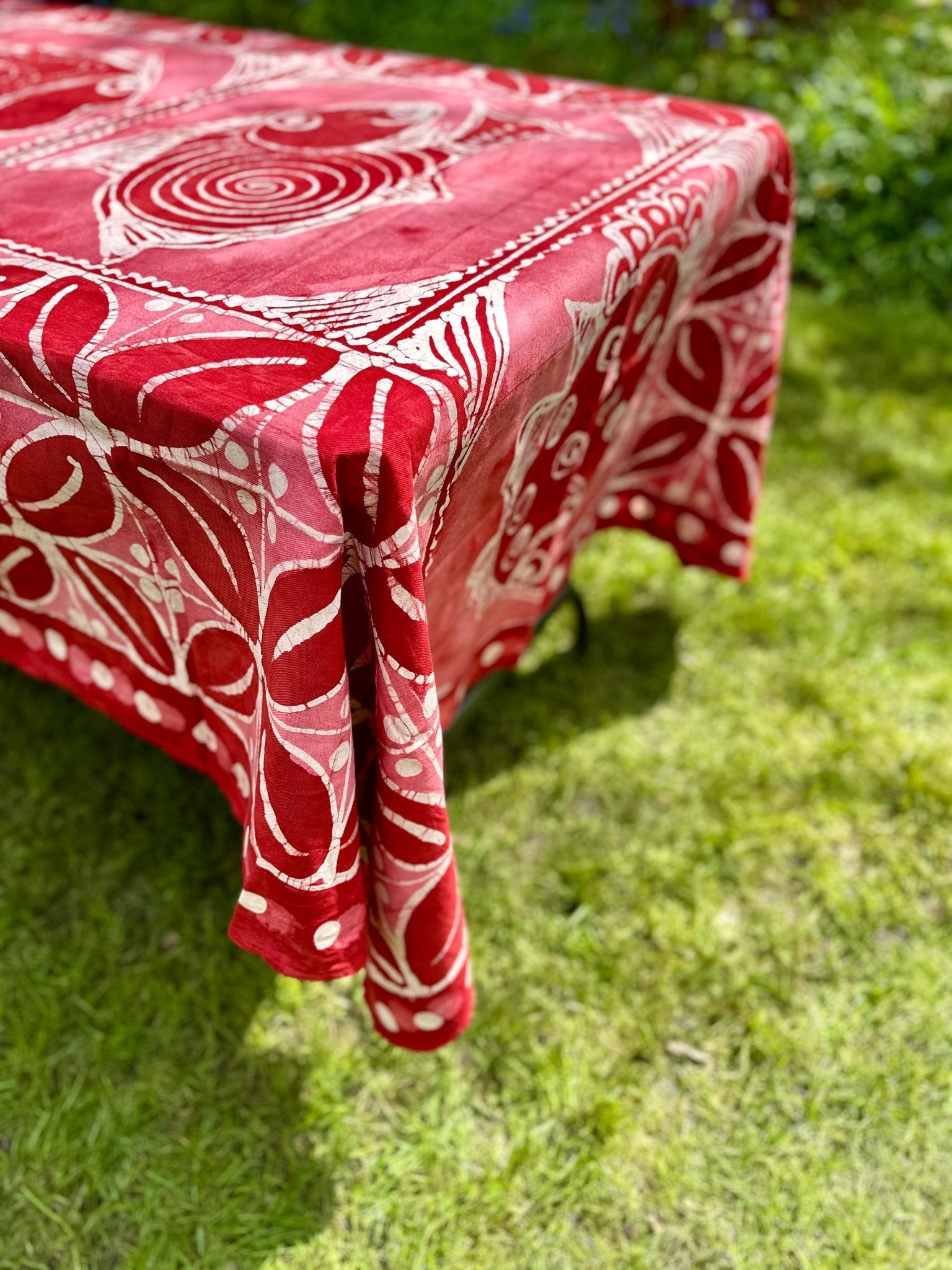 The bibianna.co.uk Batik Tablecloth / Blanket - Azų Extra Large, featuring white abstract patterns, is draped over a rectangular table set outdoors on green grass with blurred foliage in the background.
