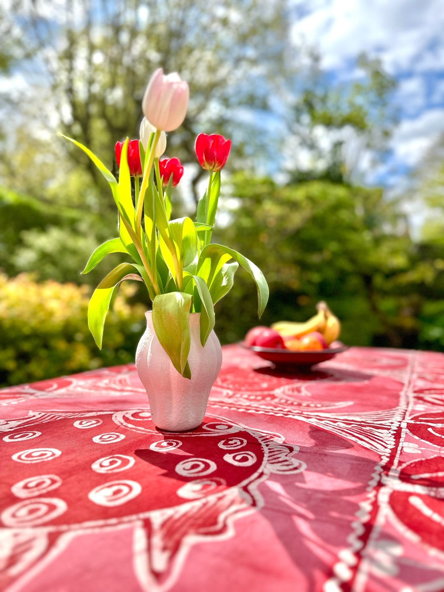 A white vase with red and white tulips sits on the bibianna.co.uk Batik Tablecloth / Blanket - Azų Extra large outdoors; a fruit bowl and green trees appear in the background beneath a partly cloudy sky.
