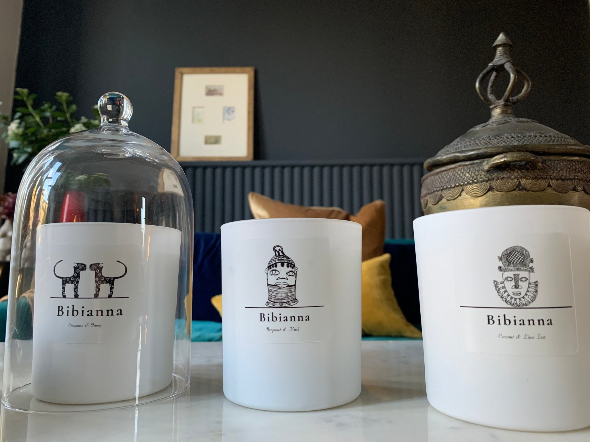 Three white Bibianaa candles with black illustrated labels sit on a marble surface; one is under a glass cloche, another is uncovered, and the third has an ornate metal lid. Colorful cushions and wall art are visible in the background.