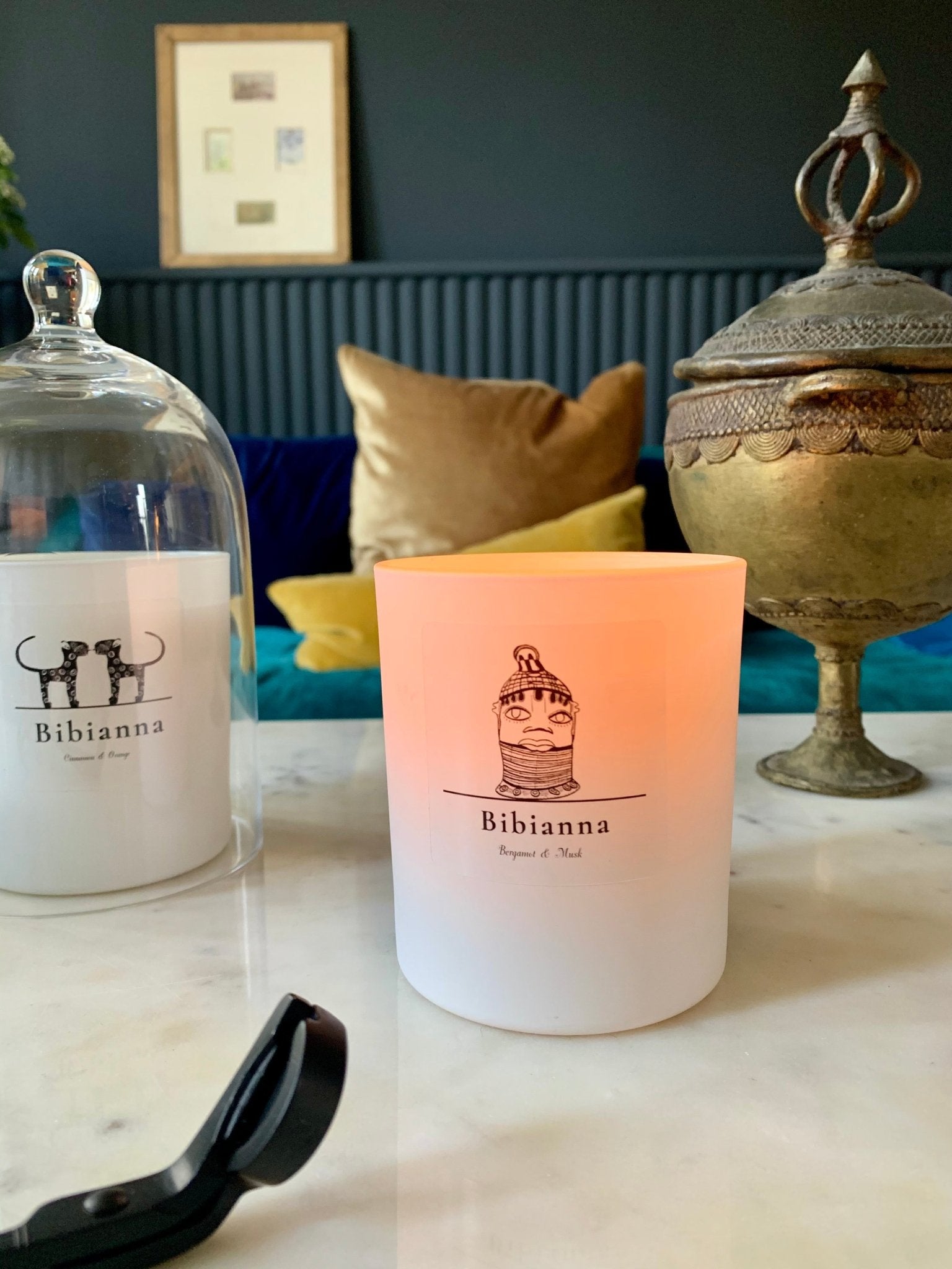 A lit Bibianna Visual Scented Candle - Bergamot & Musk by bibianna.co.uk sits on marble beside an unlit candle under a glass cloche, a gold decorative container, and cushions on a blue sofa in the background.