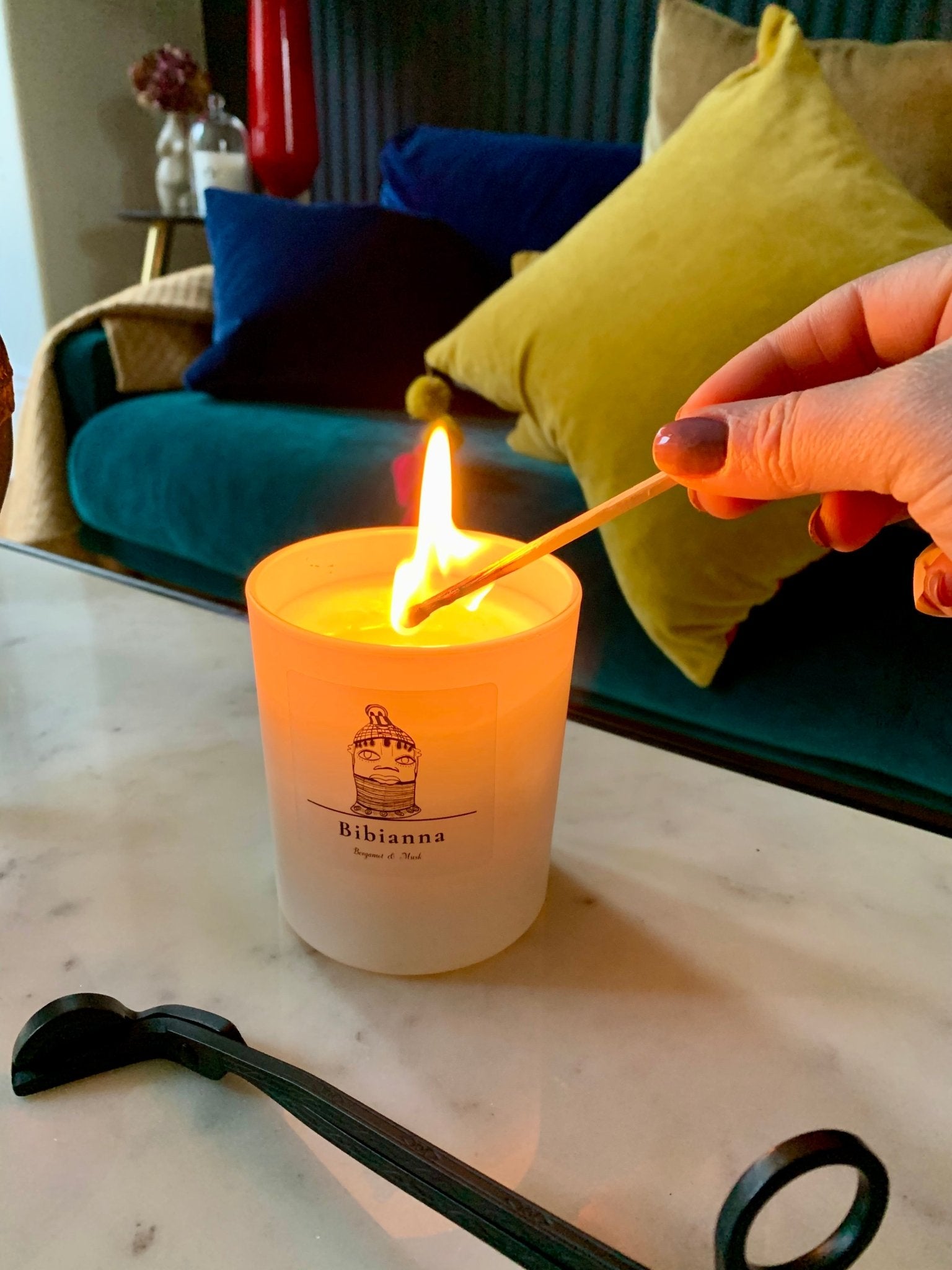 A hand lights the Bibianna Visual Scented Candle - Bergamot & Musk from bibianna.co.uk with a matchstick on a marble table, candle tools nearby and colorful pillows on a couch adding to the elevated home fragrance scene.