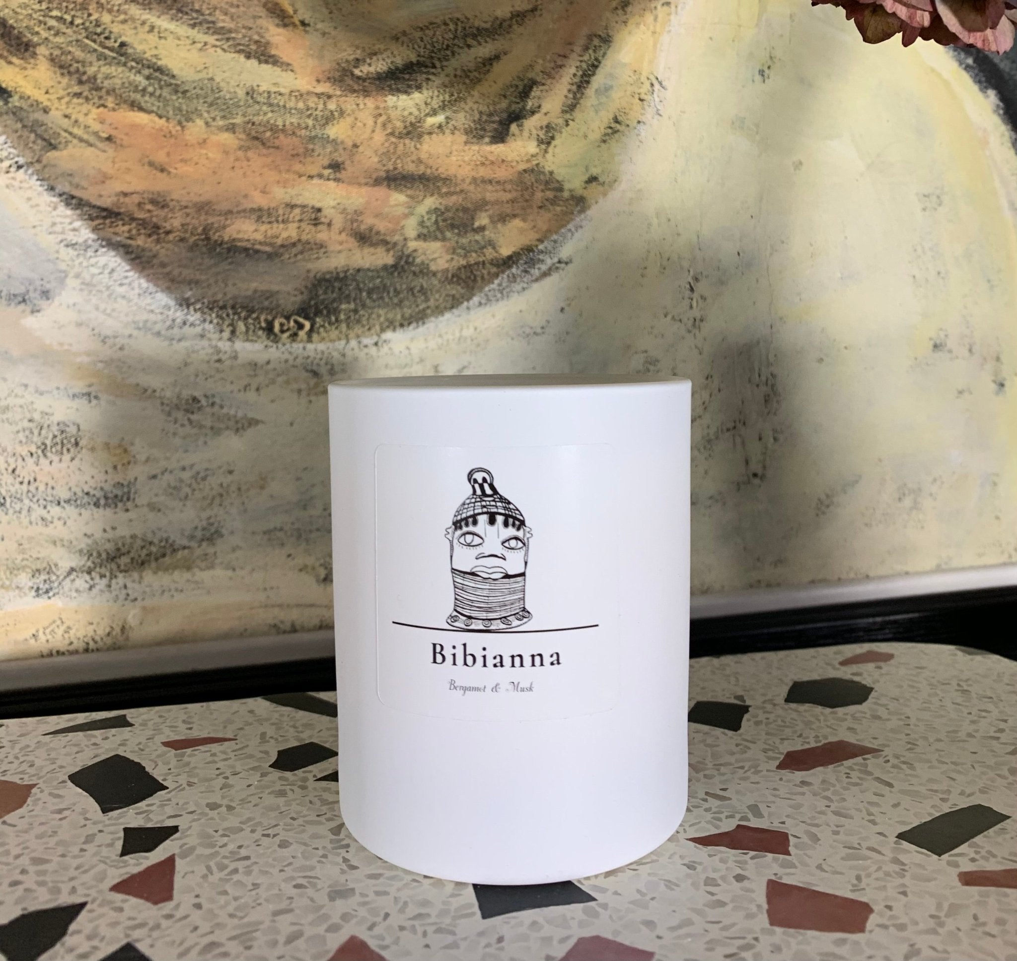 The Bibianna Visual Scented Candle – Bergamot & Musk by bibianna.co.uk features a black line drawing of a woman's face and "Bibianna" on the label, filling your room with elegant fragrance as it sits on a multicolored terrazzo surface beside an abstract painting.