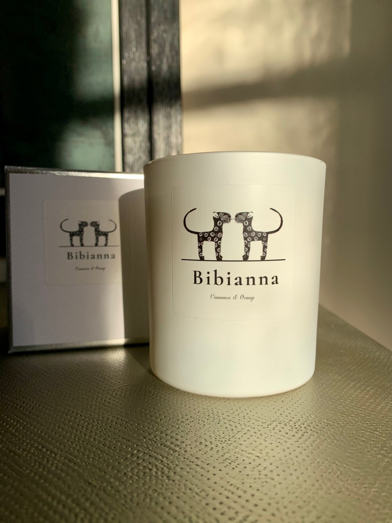 The Bibianna Luxury Scented Candle by bibianna.co.uk, with cinnamon & orange notes and twin leopard design, sits beside its box in sunlight—an elegant visual fragrance for your home collection.