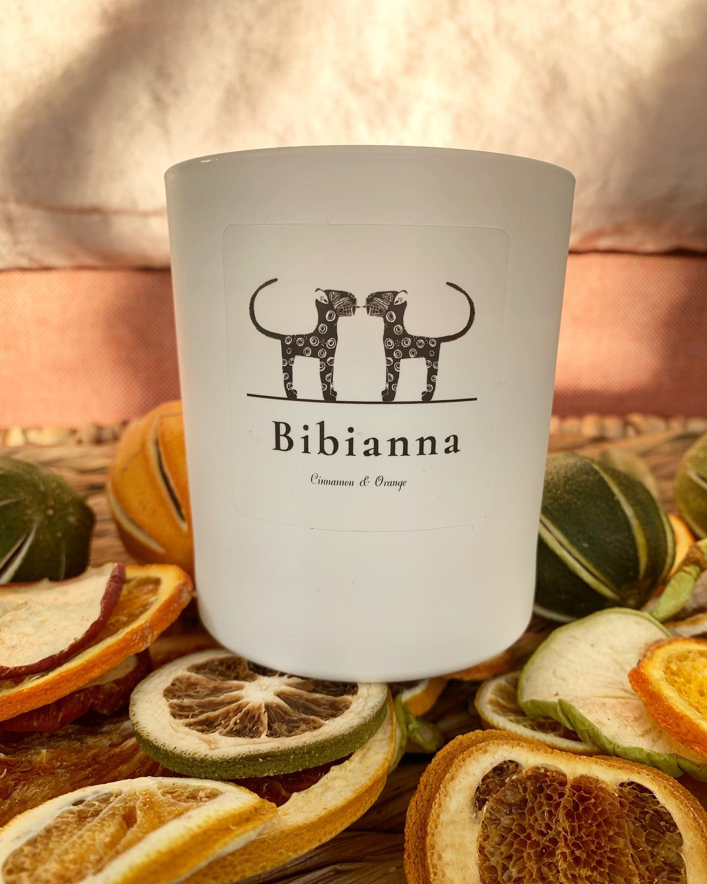 The Bibianna Luxury Scented Candles, Cinnamon & Orange by bibianna.co.uk features two cheetahs on the label, surrounded by dried citrus slices on a woven surface and bathed in warm sunlight for an inviting scene.