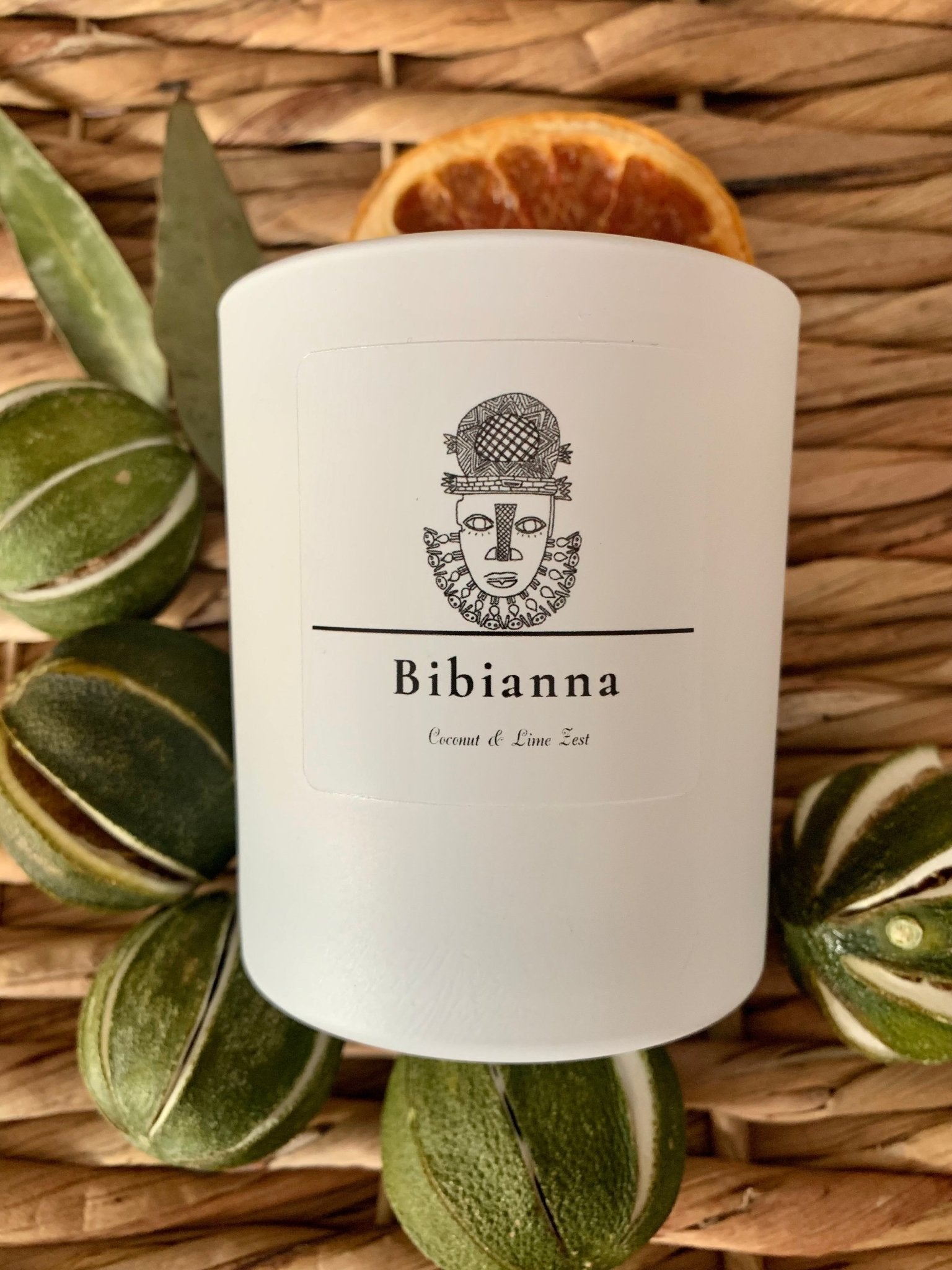 The Bibianna Scented Candle - Coconut & Lime by bibianna.co.uk rests on woven material, surrounded by green dried citrus, bay leaves, and a dried orange slice—elevate your space with this luxury hand-poured candle.