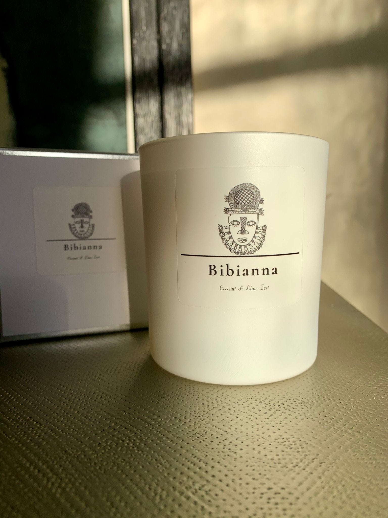 The Bibianna Scented Candle - Coconut & Lime from bibianna.co.uk features an artistic face design and comes in a matching box, both glowing in natural sunlight—a distinctive choice among luxury hand-poured candles.