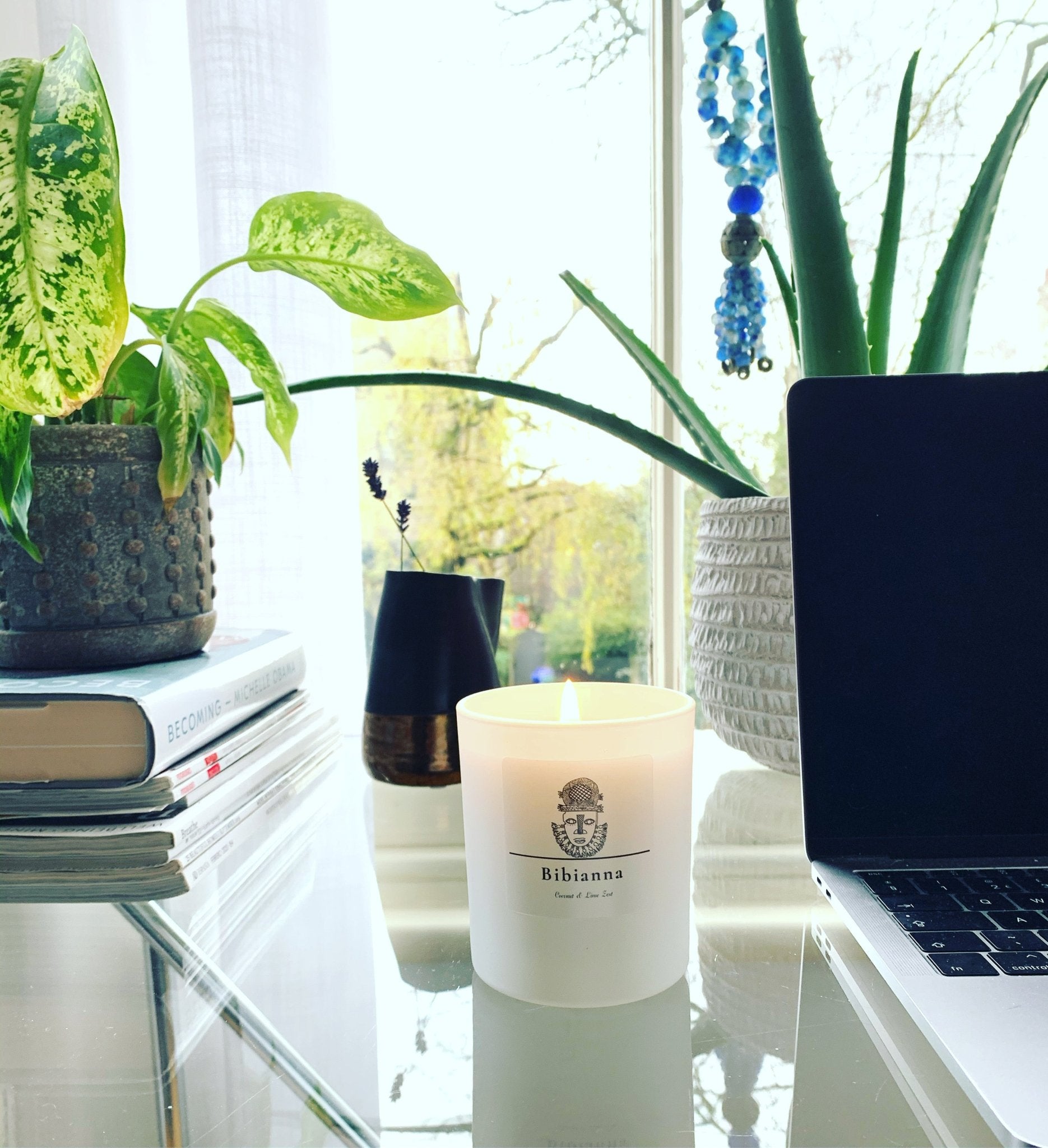 A Bibianna Scented Candle - Coconut & Lime by bibianna.co.uk sits on a glass desk near a laptop, plants, books, and blue beads, with daylight streaming in—a luxurious hand-poured home fragrance.