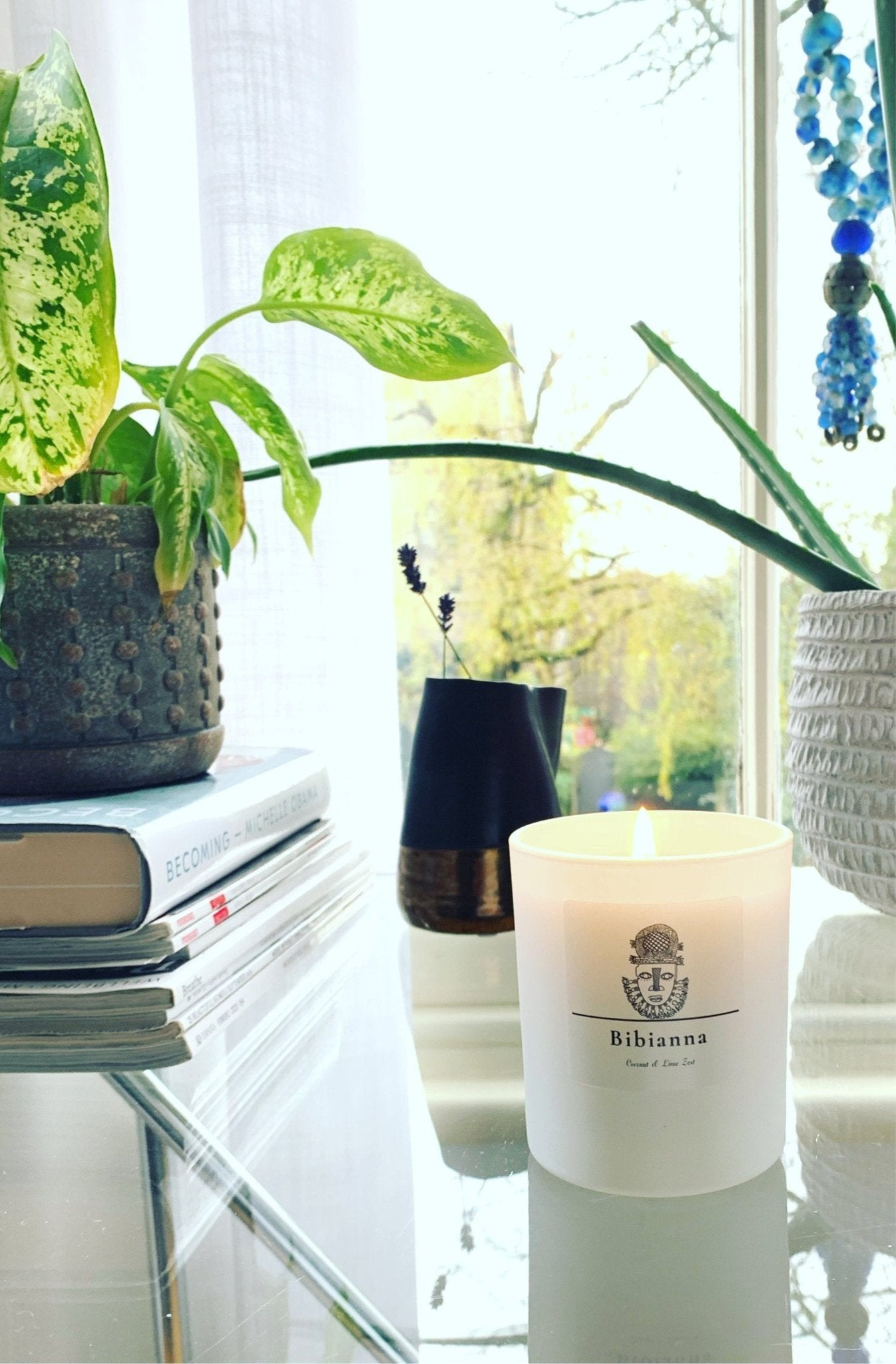 A lit Bibianna Scented Candle - Coconut & Lime from bibianna.co.uk sits on a glass table with potted plants, books, and lavender in a vase as sunlight filters through the window, enhancing its inviting fragrance.