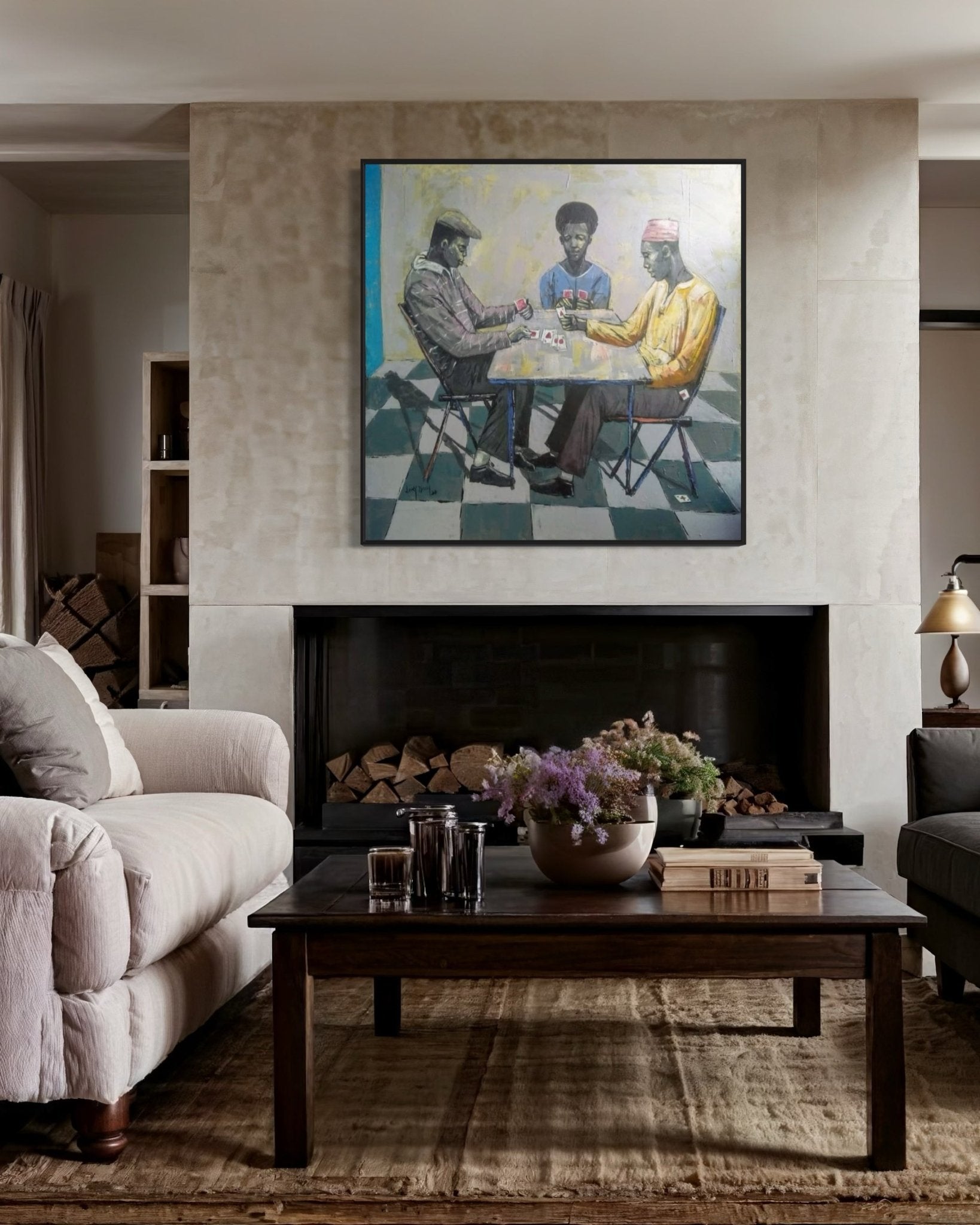 A cozy living room features a cream sofa, dark wood coffee table with flowers, log-stacked fireplace, and above it hangs "Cartels," a hand-signed Lucky Isaiah painting depicting three people sitting around a table.