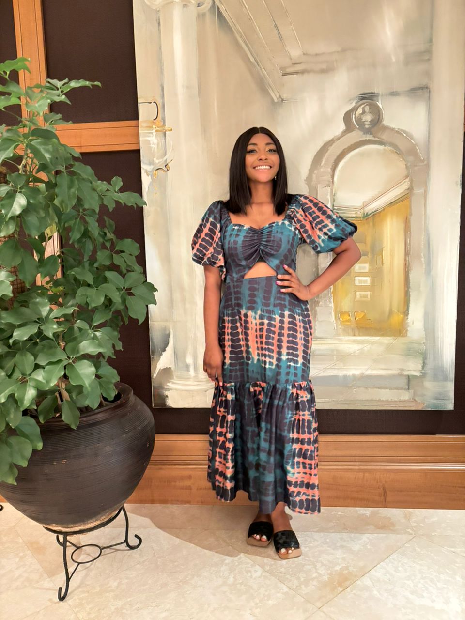 A woman in the vibrant Catalina Print Dress from bibianna.co.uk and black sandals stands indoors, smiling with one hand on her hip beside a large potted plant and a painting with architectural features.