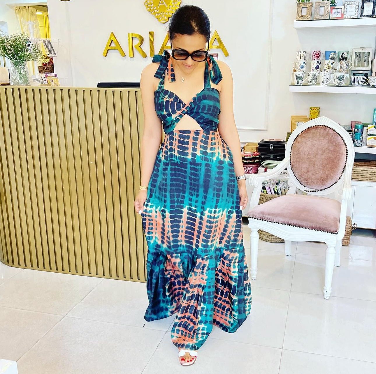 A woman in the blue and orange Catalina Print Dress from bibianna.co.uk, ideal resort wear, stands indoors with dark sunglasses and white sandals beside a gold-striped counter, a white chair with pink cushion, and display shelves.