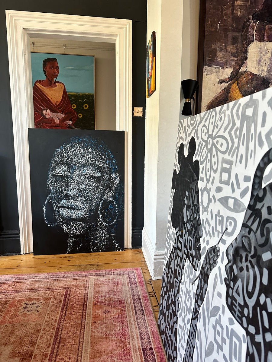 A hallway with wooden floors and a patterned rug showcases Bamidele's "Circle of Purity" artwork—a hand-signed black-and-white geometric silhouette—plus a portrait of a woman in orange and a blue abstract face.