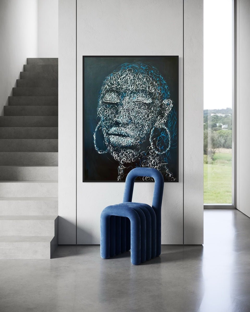 A modern interior features a blue velvet chair, curved legs, gray concrete staircase, and "Circle of purity" by Bamidele—a hand-signed abstract face portrait—on the wall. Natural light floods in through tall windows with views of greenery.