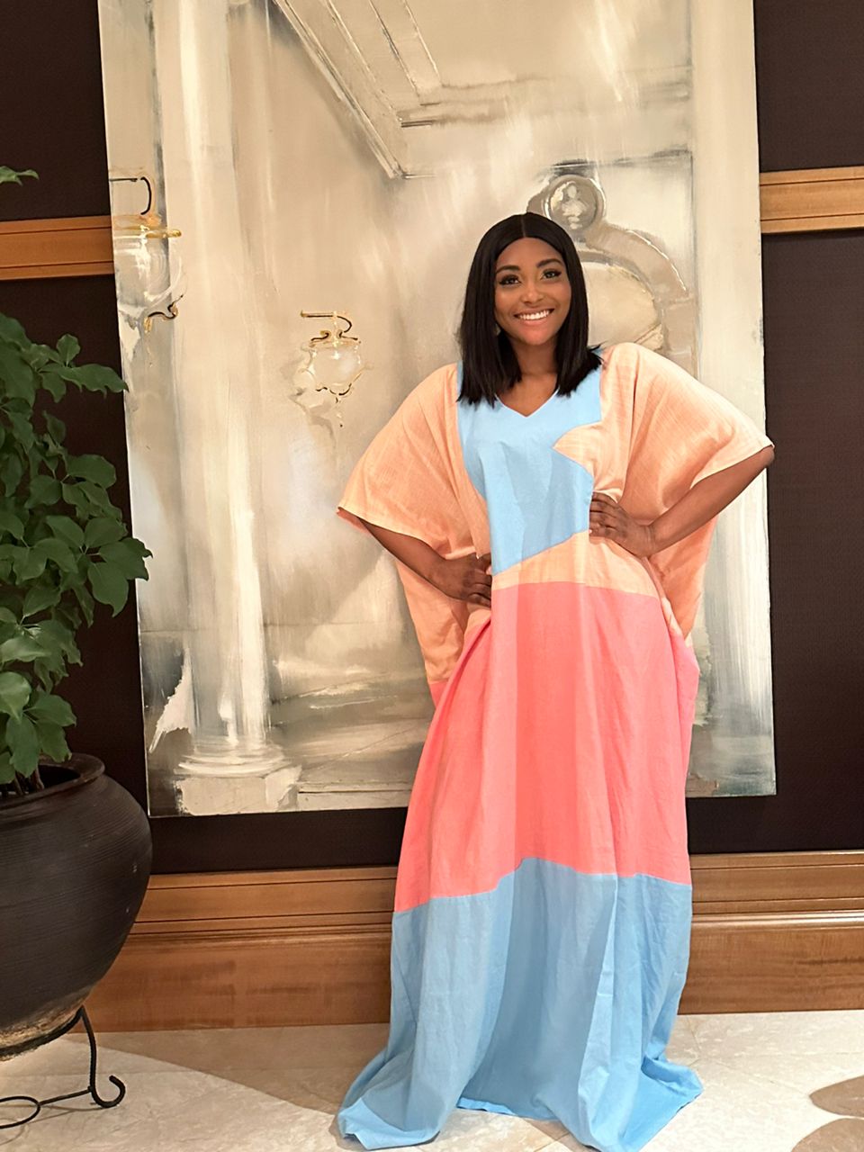 A woman wearing the Colour Block Linen Kaftan by bibianna.co.uk, featuring peach, pink, and blue panels, smiles in front of an abstract painting with her hand on her hip; a potted plant sits nearby.