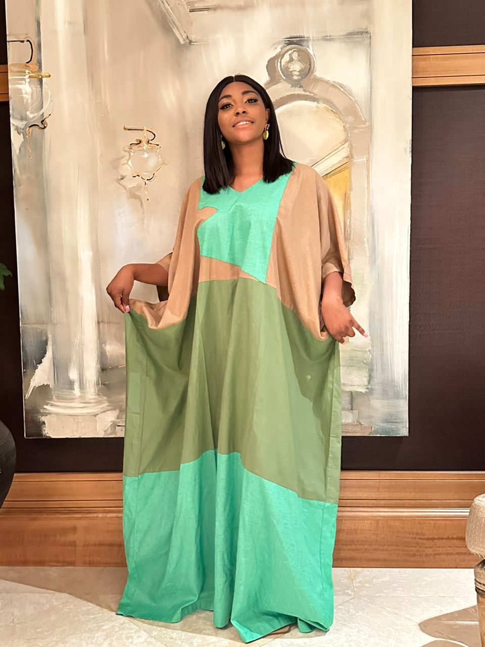 A woman stands indoors in front of an abstract painting, wearing the Colour Block Linen Kaftan Green by bibianna.co.uk, featuring green, tan, and turquoise panels. She holds the sides of her dress and smiles at the camera.