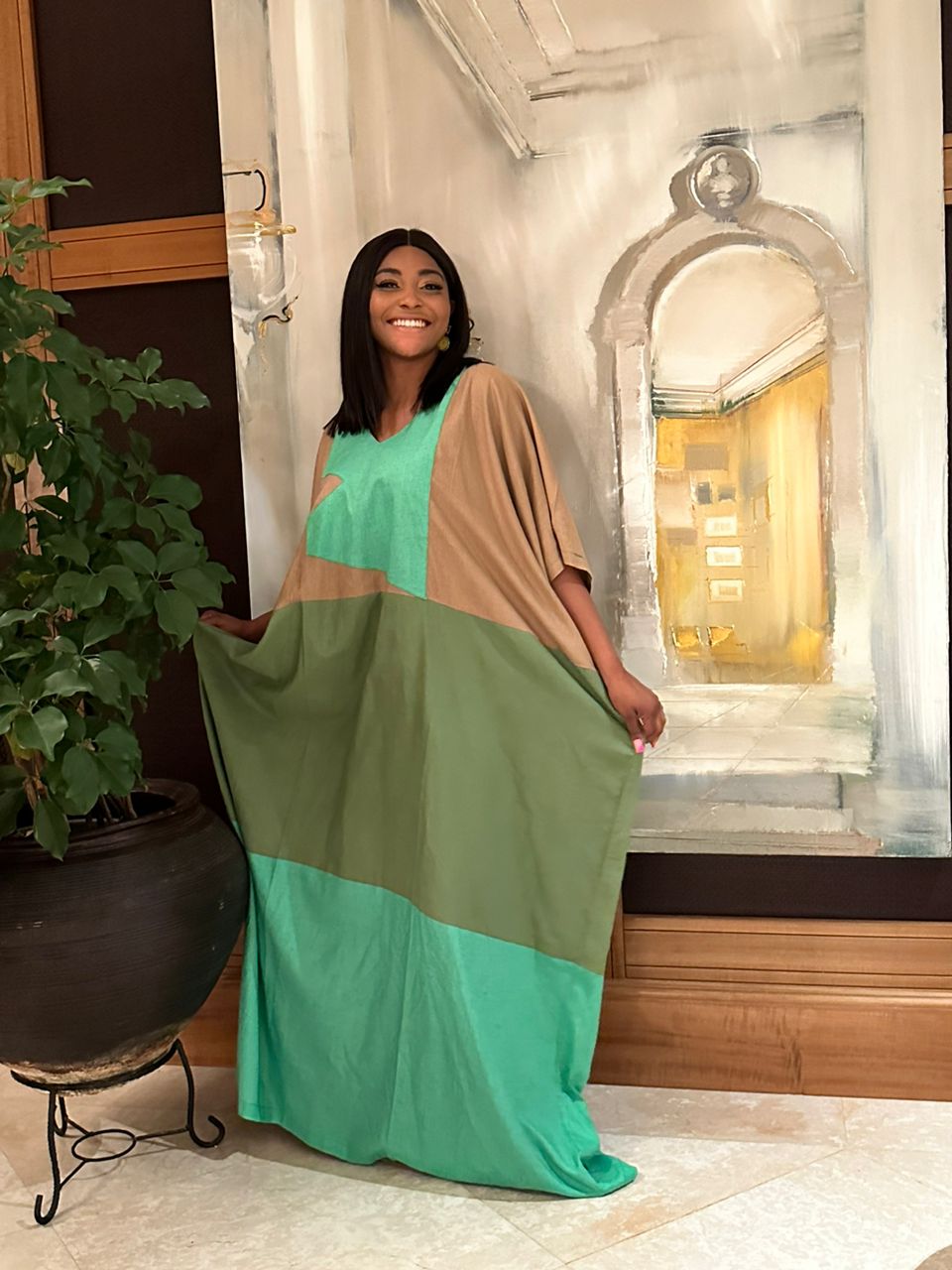 A woman smiles in front of a large arched painting, wearing the Colour Block Linen Kaftan Green from bibianna.co.uk, with a potted plant beside her.