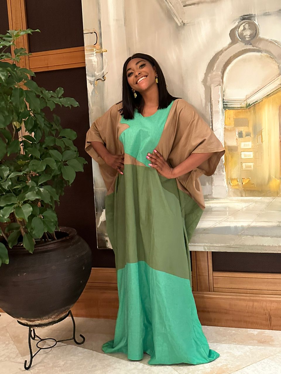 A smiling woman models the Colour Block Linen Kaftan Green from bibianna.co.uk, standing indoors before a large painting with her hands on her hips and a potted plant to her left.