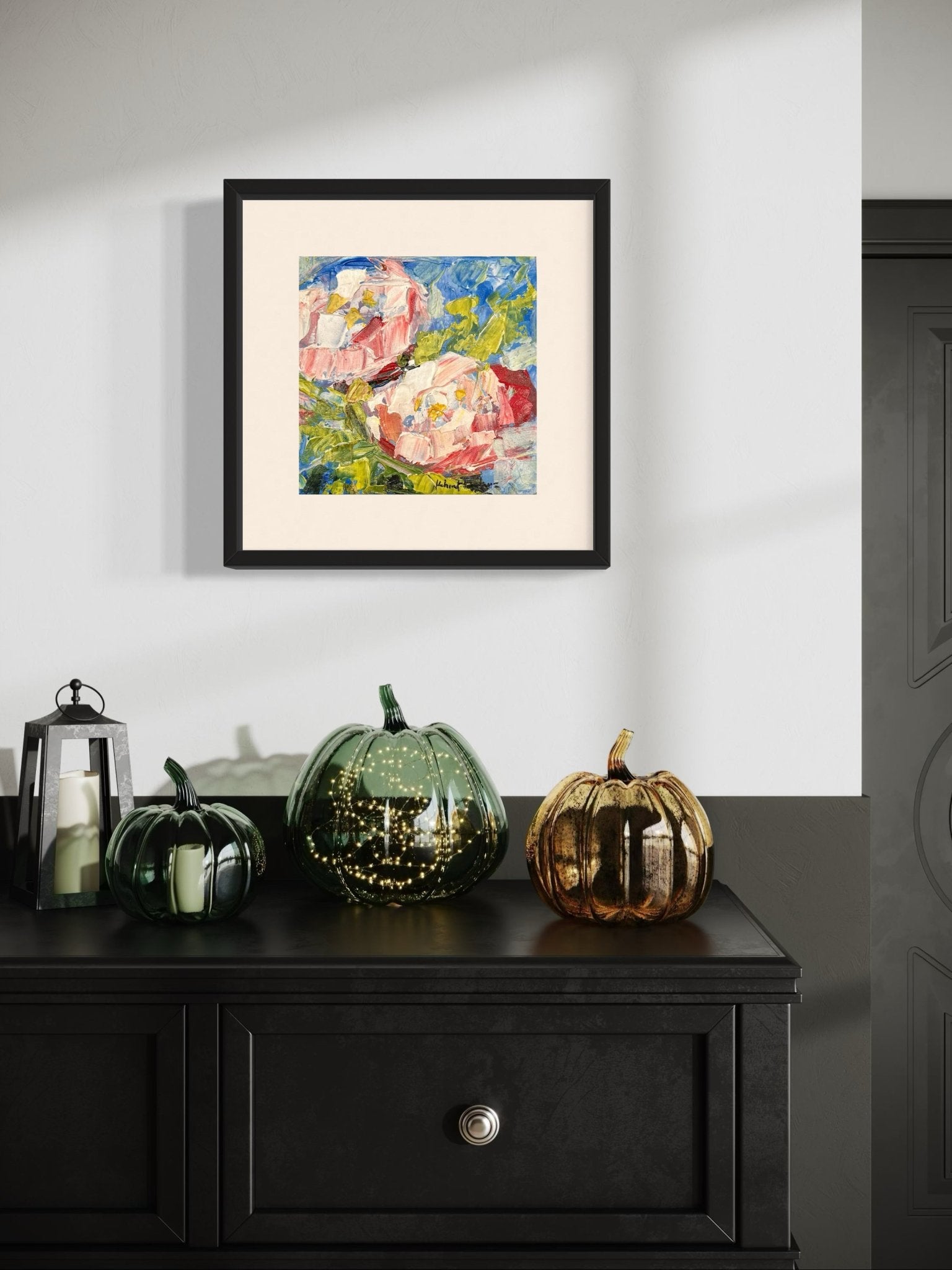 A black dresser with three decorative pumpkins and candles sits below a framed Contemporary Textured Abstract Floral Miniature Painting by Kehinde Badmuss on a white wall near a gray door, while sunlight casts soft shadows across the scene.