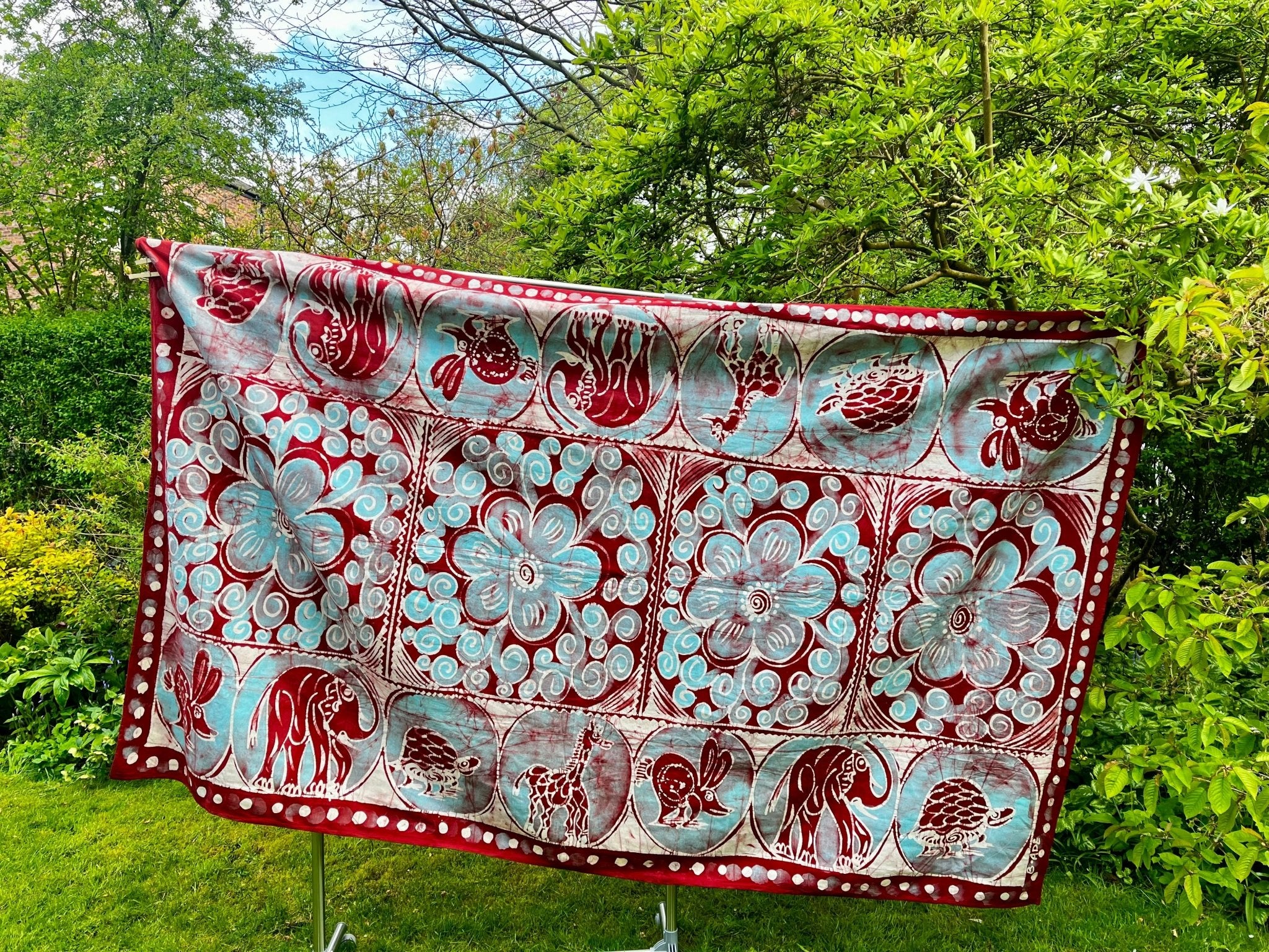 The Cotton Batik Freehand Illustrated Table Cloth - Ifuru by bibianna.co.uk, featuring vibrant red, white, and blue floral and animal designs, hangs on a rack outdoors amid lush garden greenery.