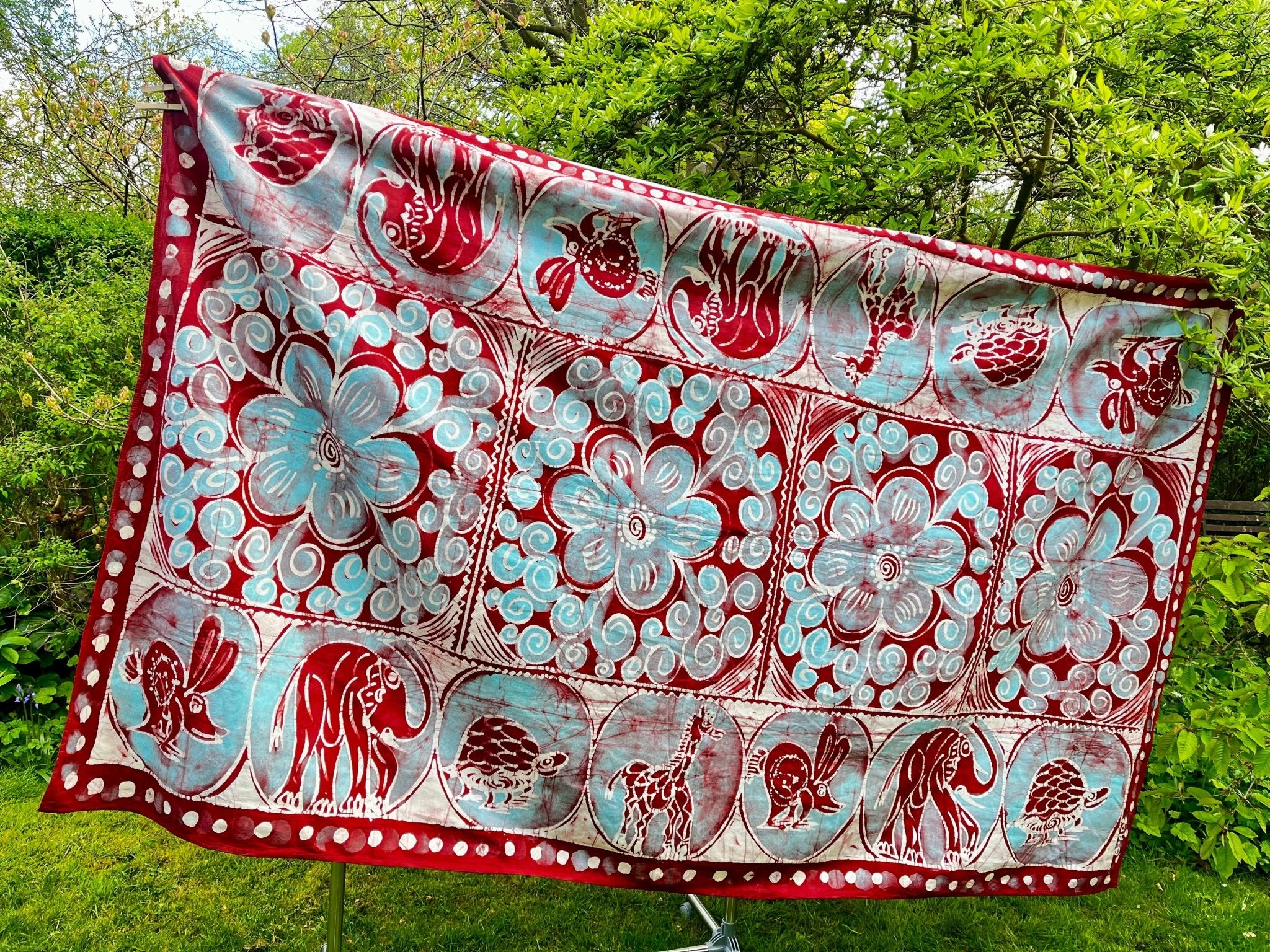 The Cotton Batik Freehand Illustrated Table Cloth - Ifuru from bibianna.co.uk, featuring vibrant red and blue floral patterns with elephants, turtles, and birds, is displayed outside against green trees and grass.