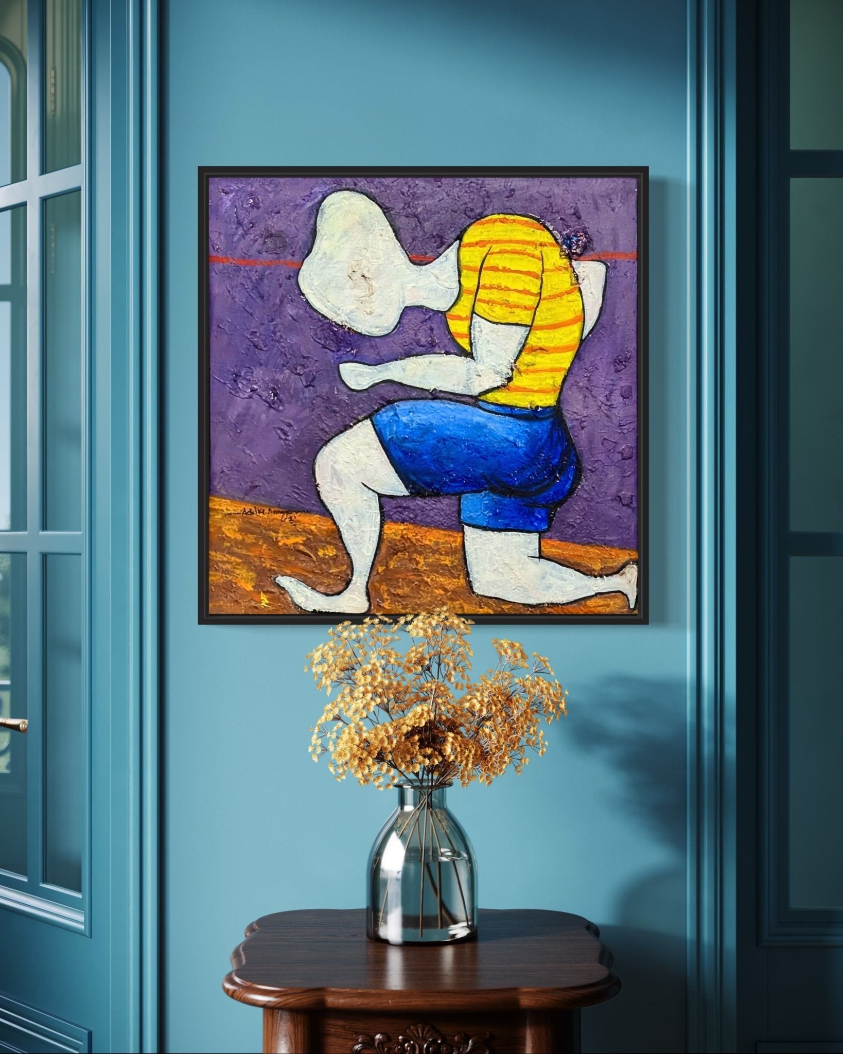 Dance Of Nature" by Achike Anayo, an expressionist painting of an abstract figure in a yellow striped shirt and blue shorts, is hand-signed and displayed in a black tray frame on a blue wall above a wooden table with dried flowers.
