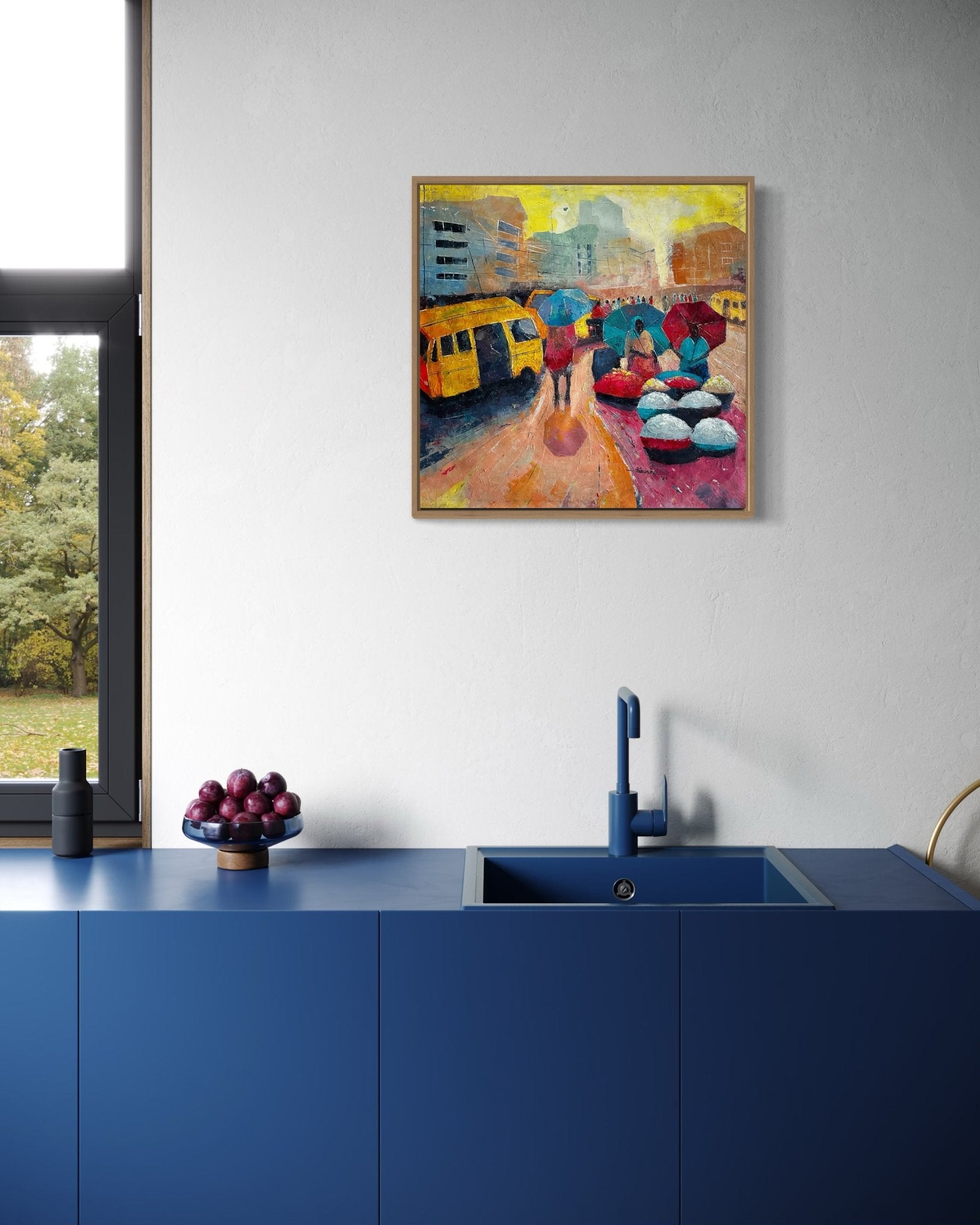 A modern kitchen with matte blue cabinets and sink, a bowl of plums on the counter, and "Danfo" by Bibianna African Art Collective—an artwork depicting Lagos street life—hanging on the white wall above.
