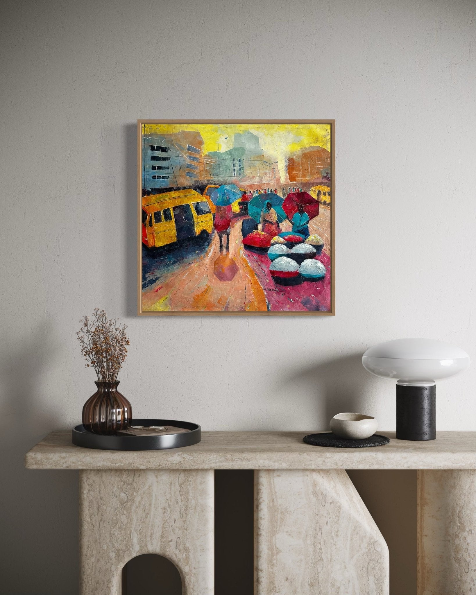 A framed "Danfo" painting by Bibianna African Art Collective—showing Lagos street life with umbrellas and a yellow danfo bus—hangs above a beige console table topped with a vase, round tray, and modern lamp.