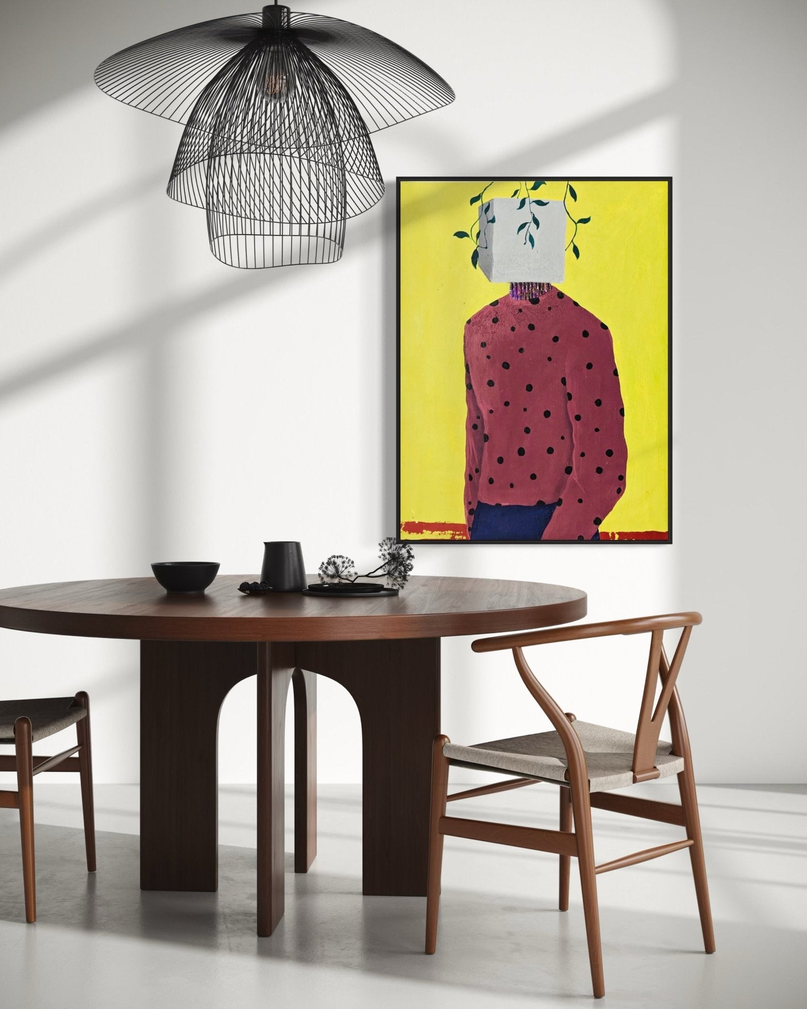 A modern dining area features a round wooden table, two chairs, and a black pendant lamp. The white wall displays "Embody II" by Slam Austin, a vibrant acrylic on canvas of a faceless figure in a polka-dot shirt against yellow.