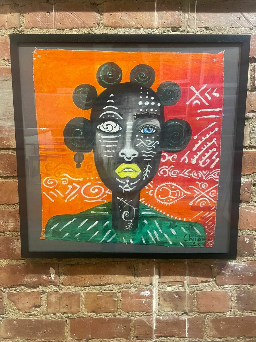 Eyes Of Symbol" by Chisom S is a framed acrylic-on-canvas artwork featuring a stylized face with green lips, geometric and tribal patterns, set against a vibrant orange and red background that stands out on any brick wall.